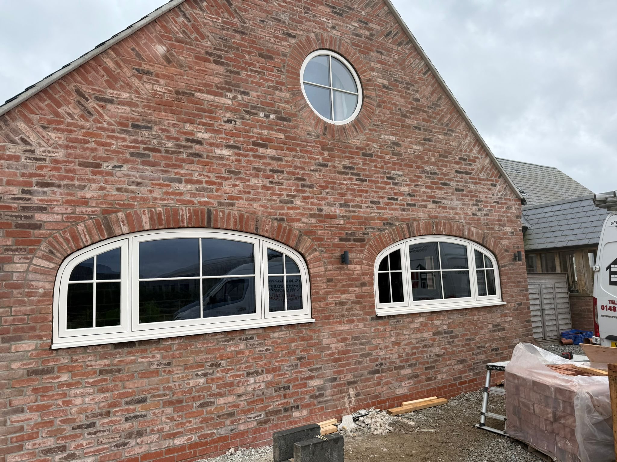 A brick house with three white-framed, arched windows, two on the bottom level and one smaller circular window above, under a cloudy sky. There is construction equipment and materials in front of the house.