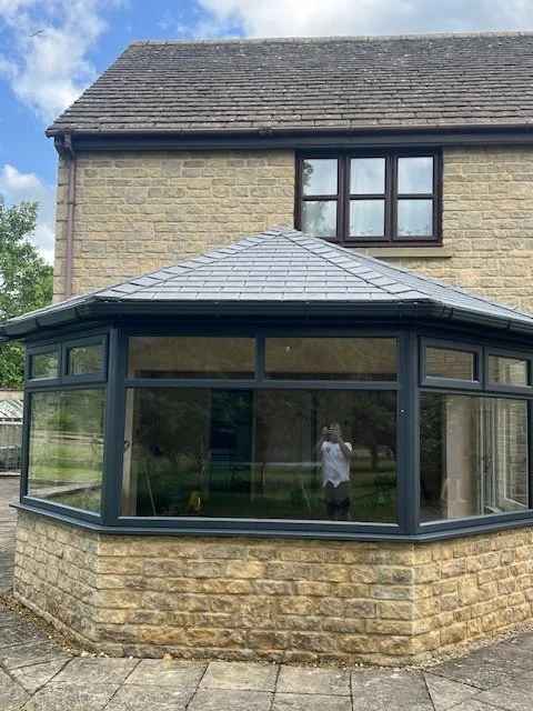 The image shows the exterior of a two-story house with beige brick walls and a window on the upper floor. Attached to the house is a basement extension with large glass windows and a tiled roof, reflecting the surrounding yard and the person taking the photo.