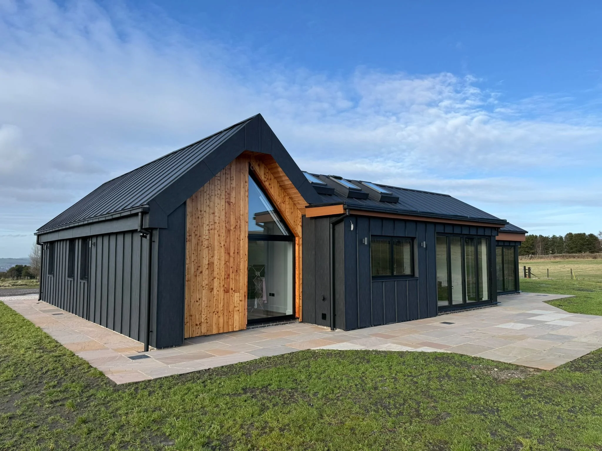 Modern black house with wooden accents and large glass windows situated on a grassy landscape under a partly cloudy sky.