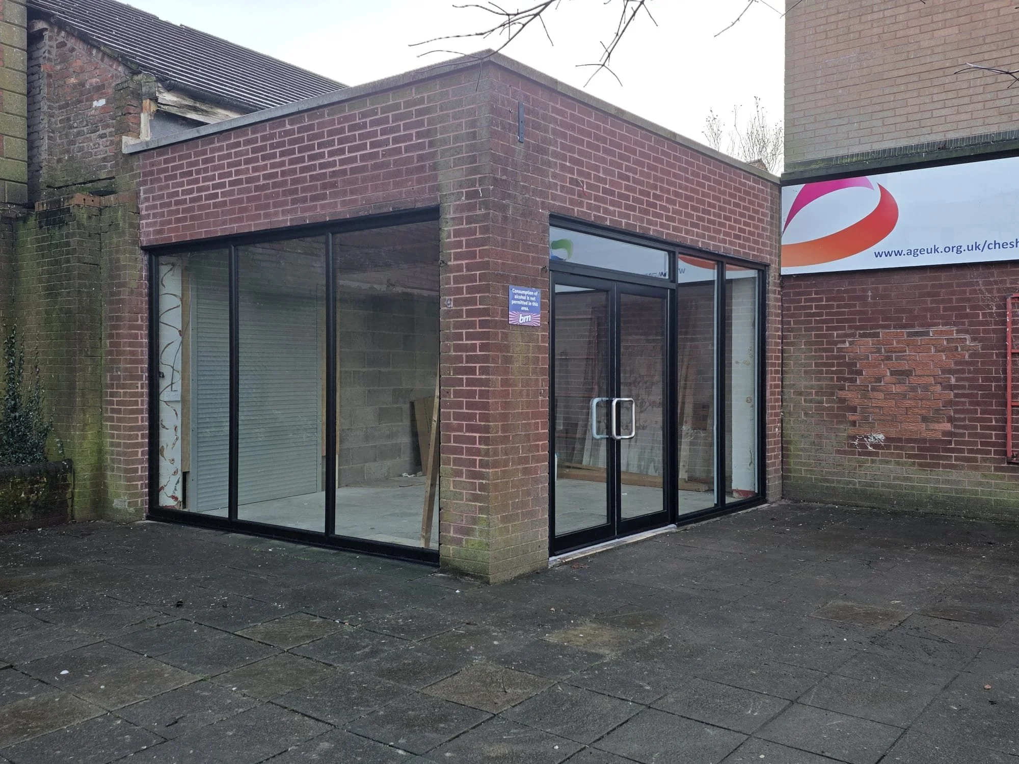 A small brick building with large glass windows and door, with an outdoor paved area in front, and a sign on the wall indicating a COVID-19 safety notice.