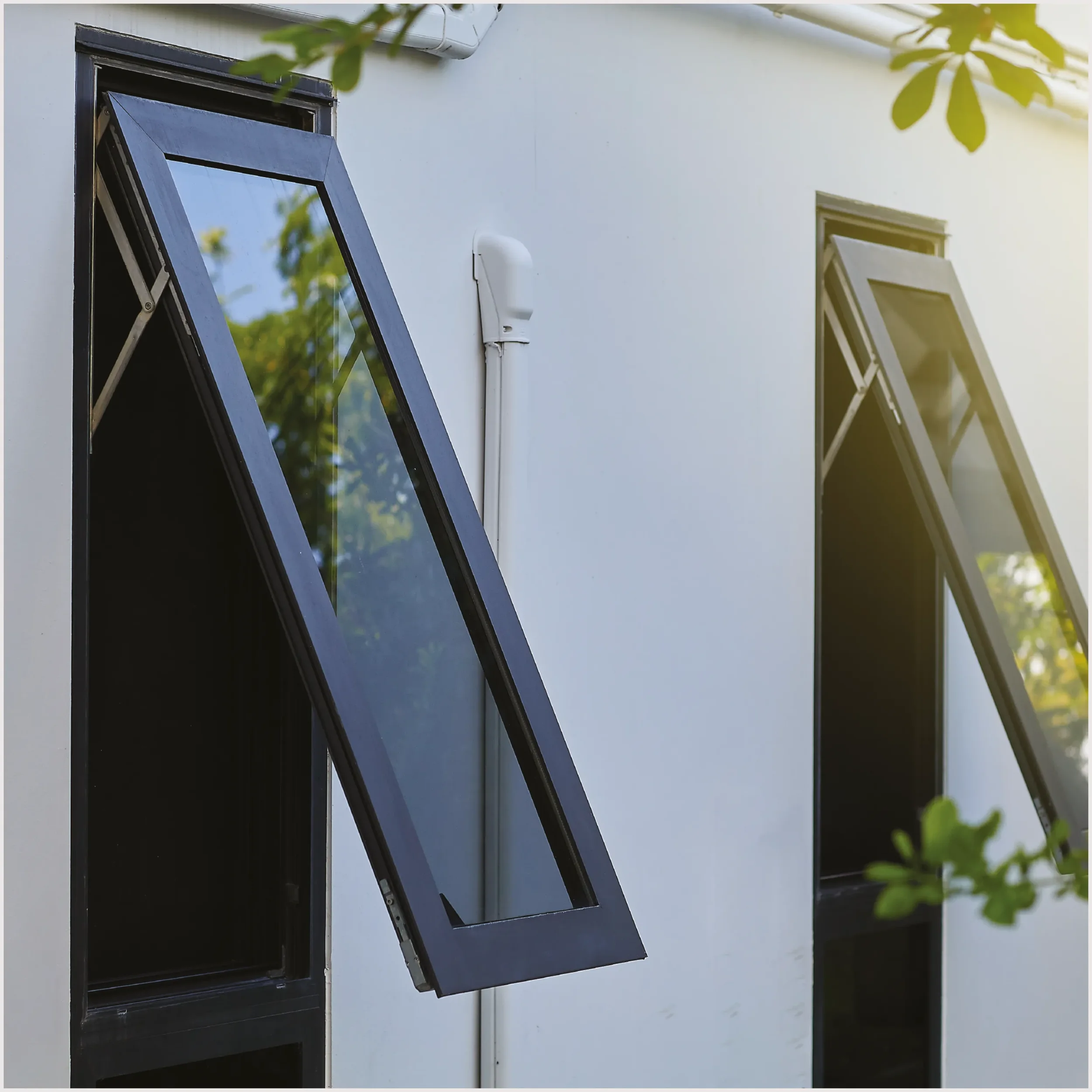 Open black window with a reflective glass pane on a white building wall, surrounded by green foliage and a blue sky reflected in the glass.