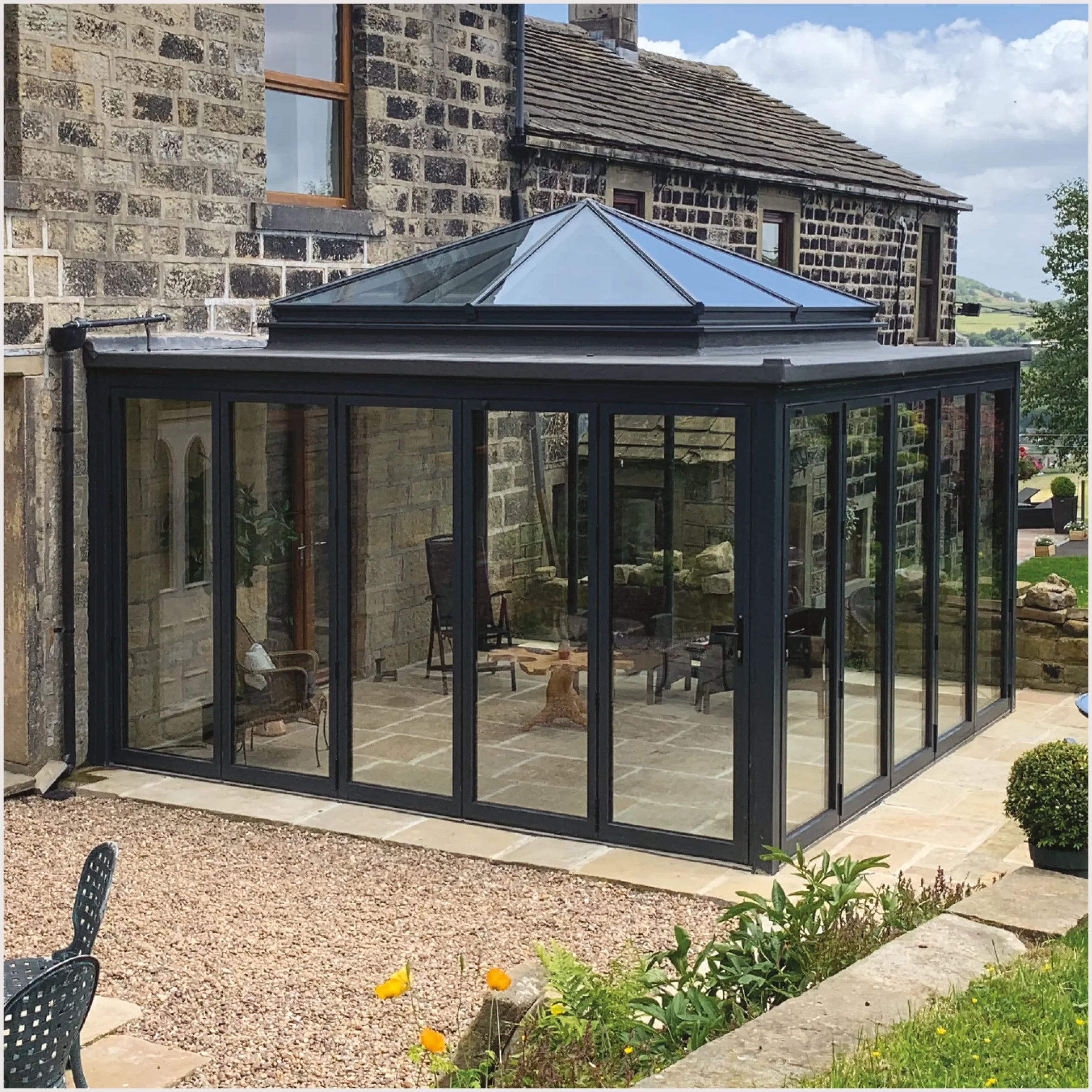 View of a backyard patio area with a stone house, a glass conservatory, and a garden with flowers and outdoor furniture.