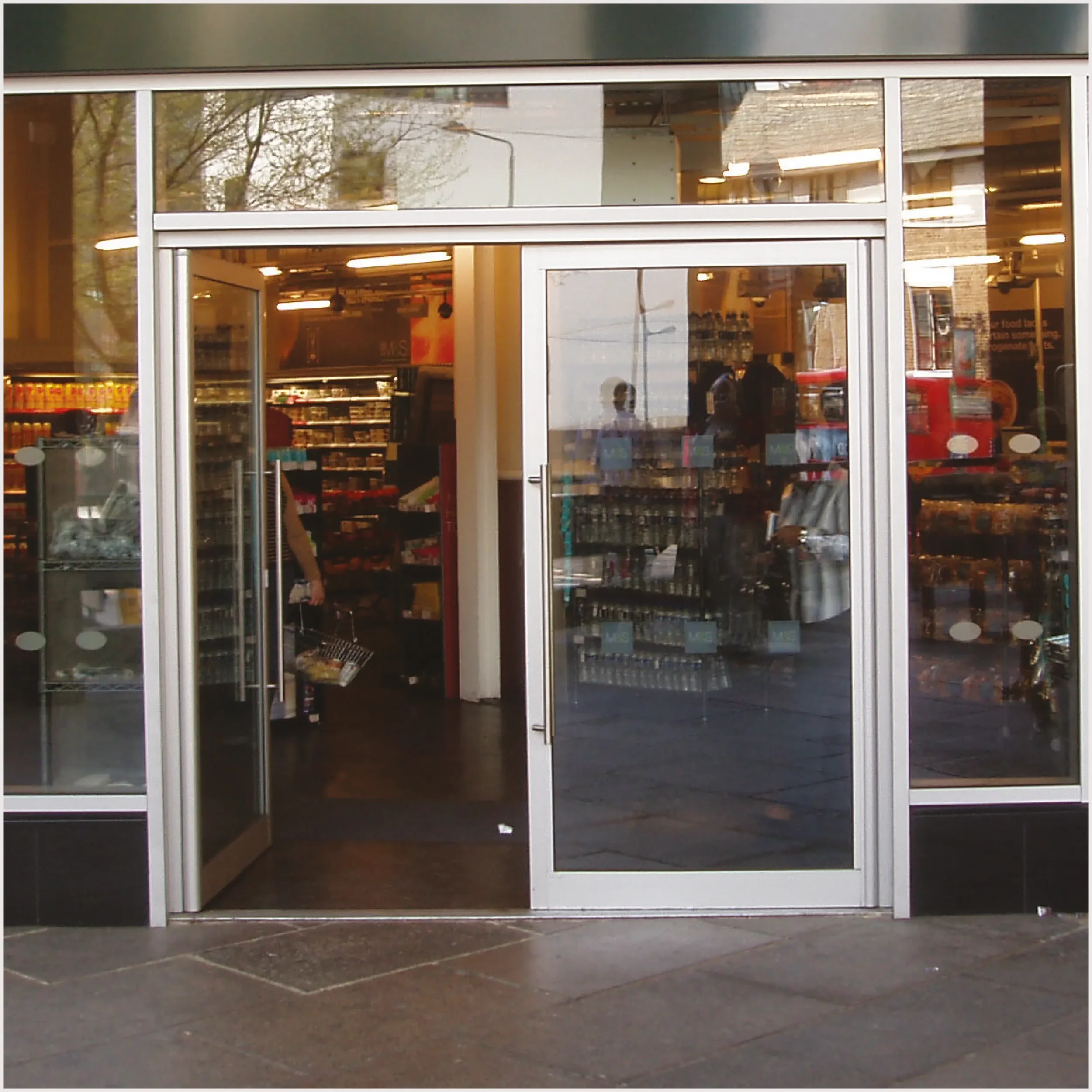 View of the entrance to a grocery store with glass doors and windows, showing the store interior and some items on shelves inside.