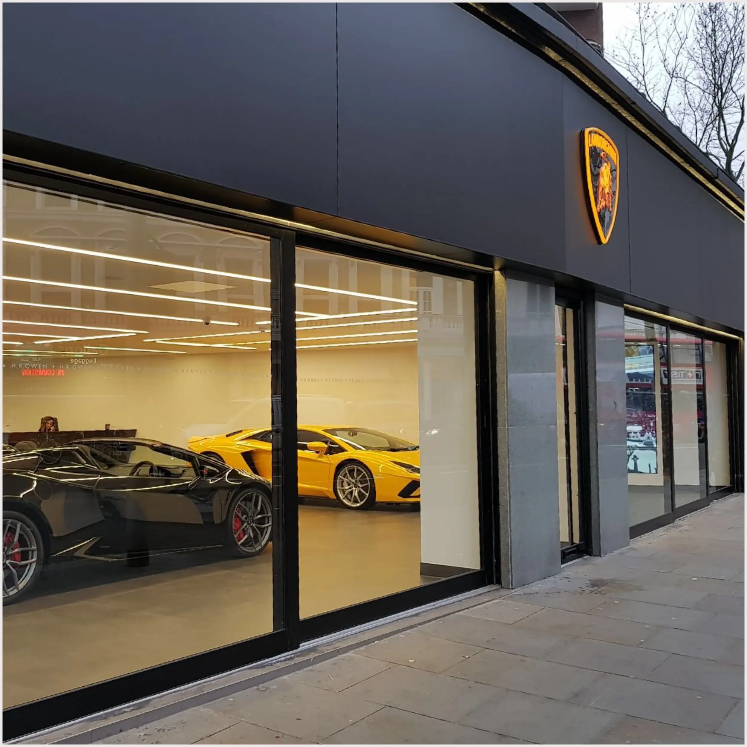 Interior of a luxury car dealership showroom with a yellow sports car visible through the window.