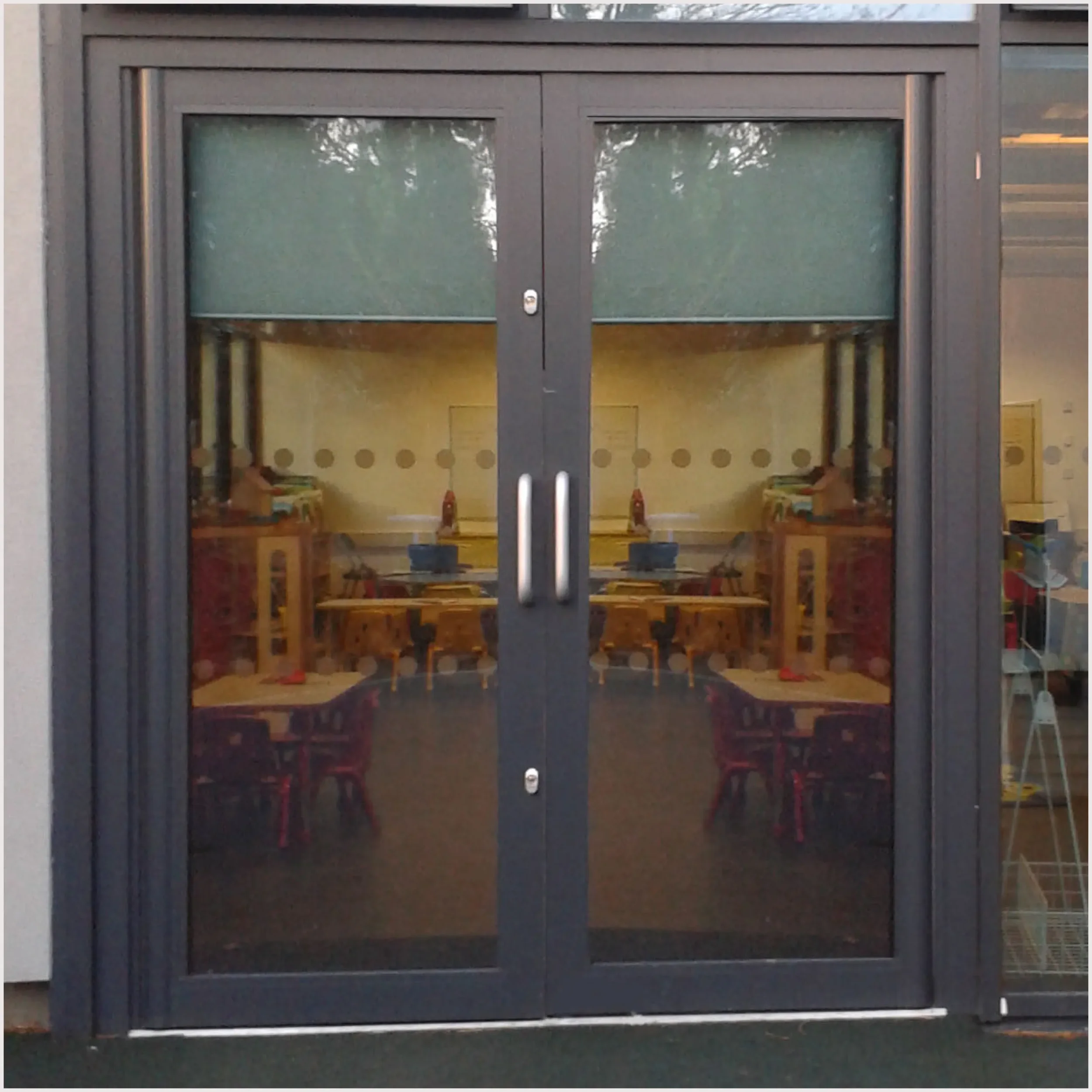 Closed glass door with dark metal frame, showing a reflection of trees in the upper glass pane, leading into a room with wooden tables and chairs.