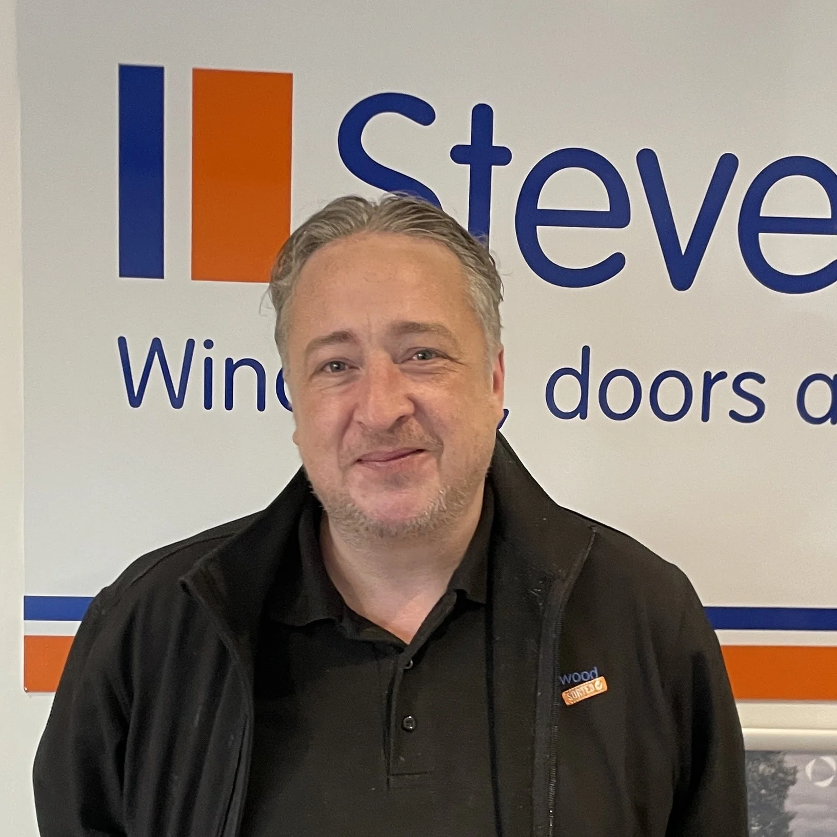 A middle-aged man smiling at the camera, standing in front of a sign that says "I believe in" with partial text visible. He is wearing a black jacket and a black shirt.