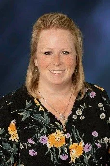 Portrait of a woman with blonde hair wearing a floral blouse, smiling, against a blue background.
