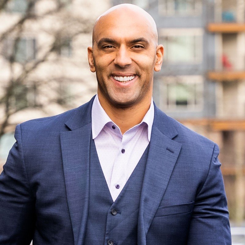 A smiling man in a navy suit and light purple shirt standing outdoors with buildings and trees in the background.