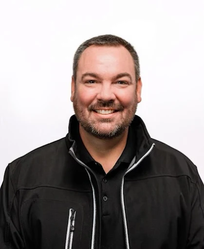 Headshot of a smiling man with a beard, wearing a black jacket and black shirt, against a white background.