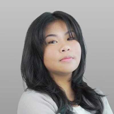 Portrait of a woman with dark hair, wearing a light-colored top, looking confidently at the camera with a neutral gray background.