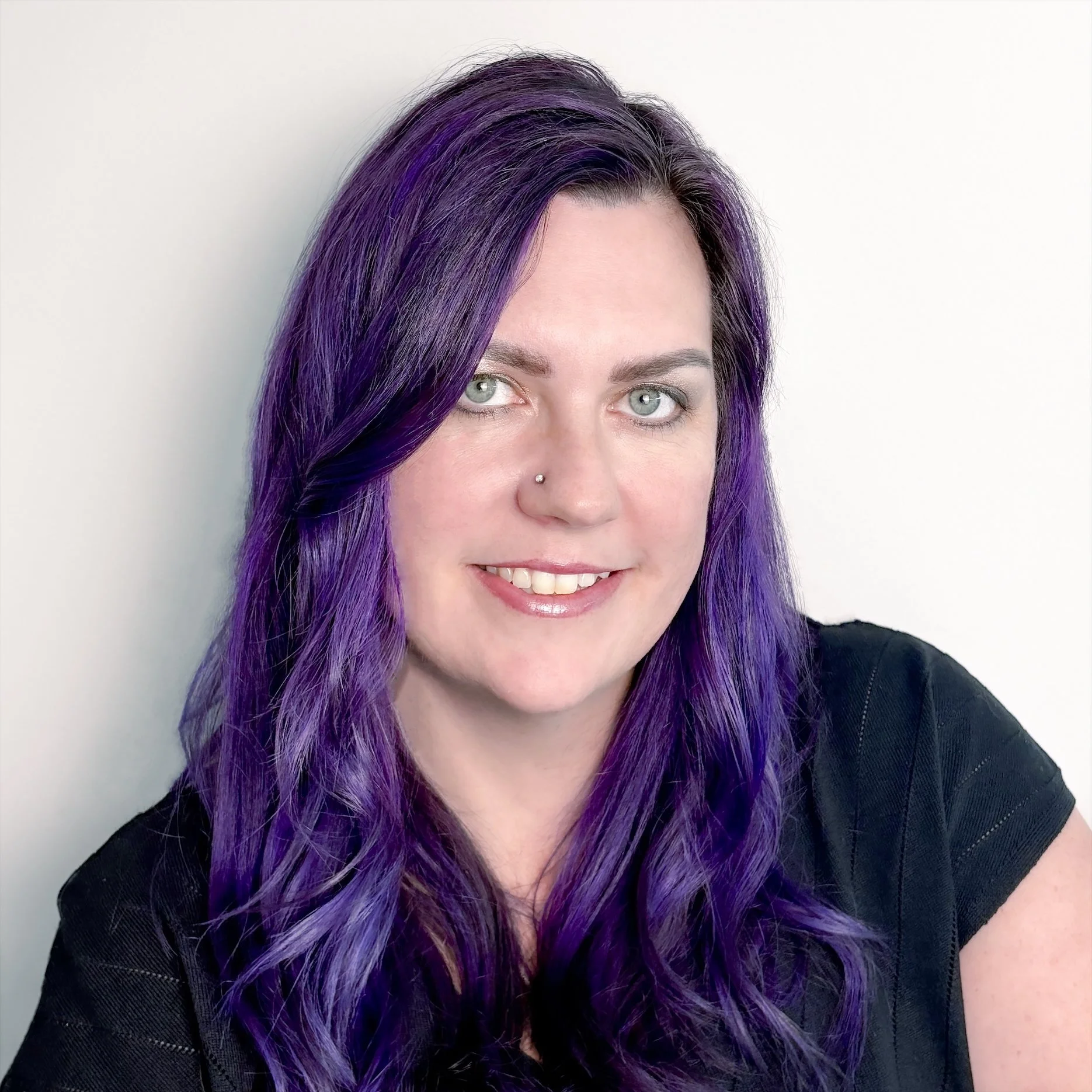 Close-up of a woman with light gray eyes and stylish purple hair, wearing a black top against a plain, light-colored background.