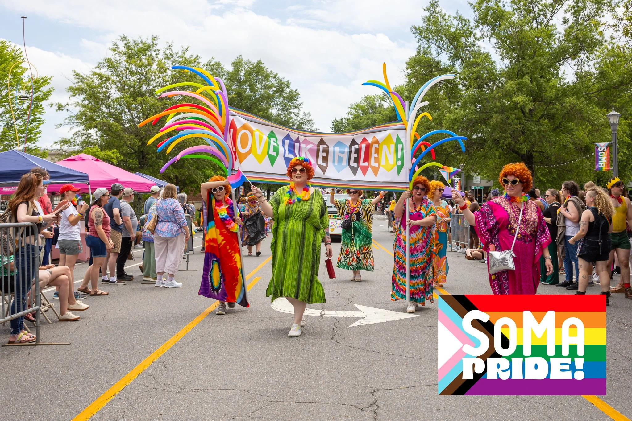 People in colorful outfits walking in a parade holding a rainbow-themed banner that says "Love Like Helen!" with rainbow balloons. Spectators watch from the sides. There is a sign saying "SOMA Pride!" in the lower right corner.