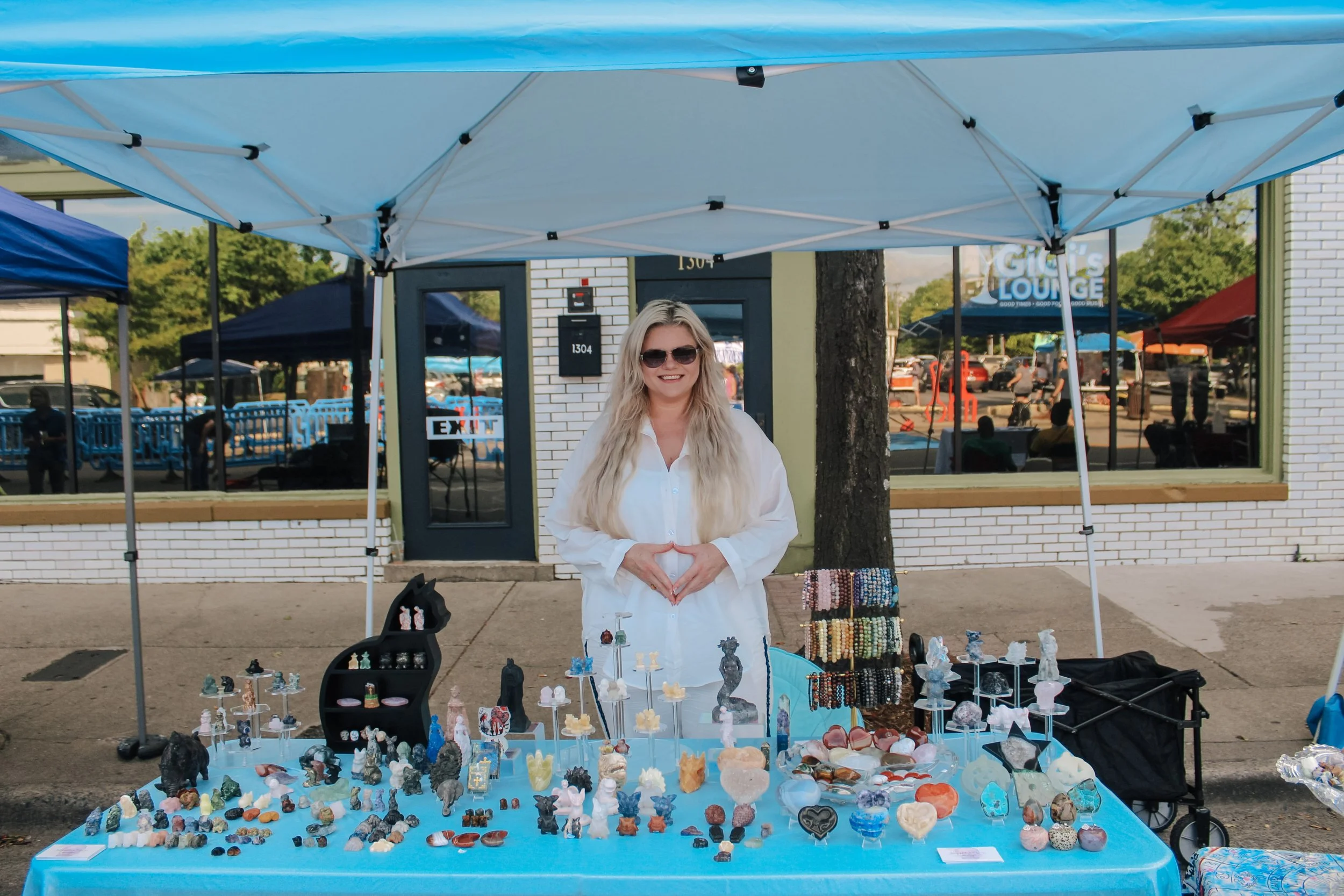Woman standing behind a table with jewelry and stone crafts at outdoor market booth, wearing sunglasses and white shirt, with tents and storefronts in background.