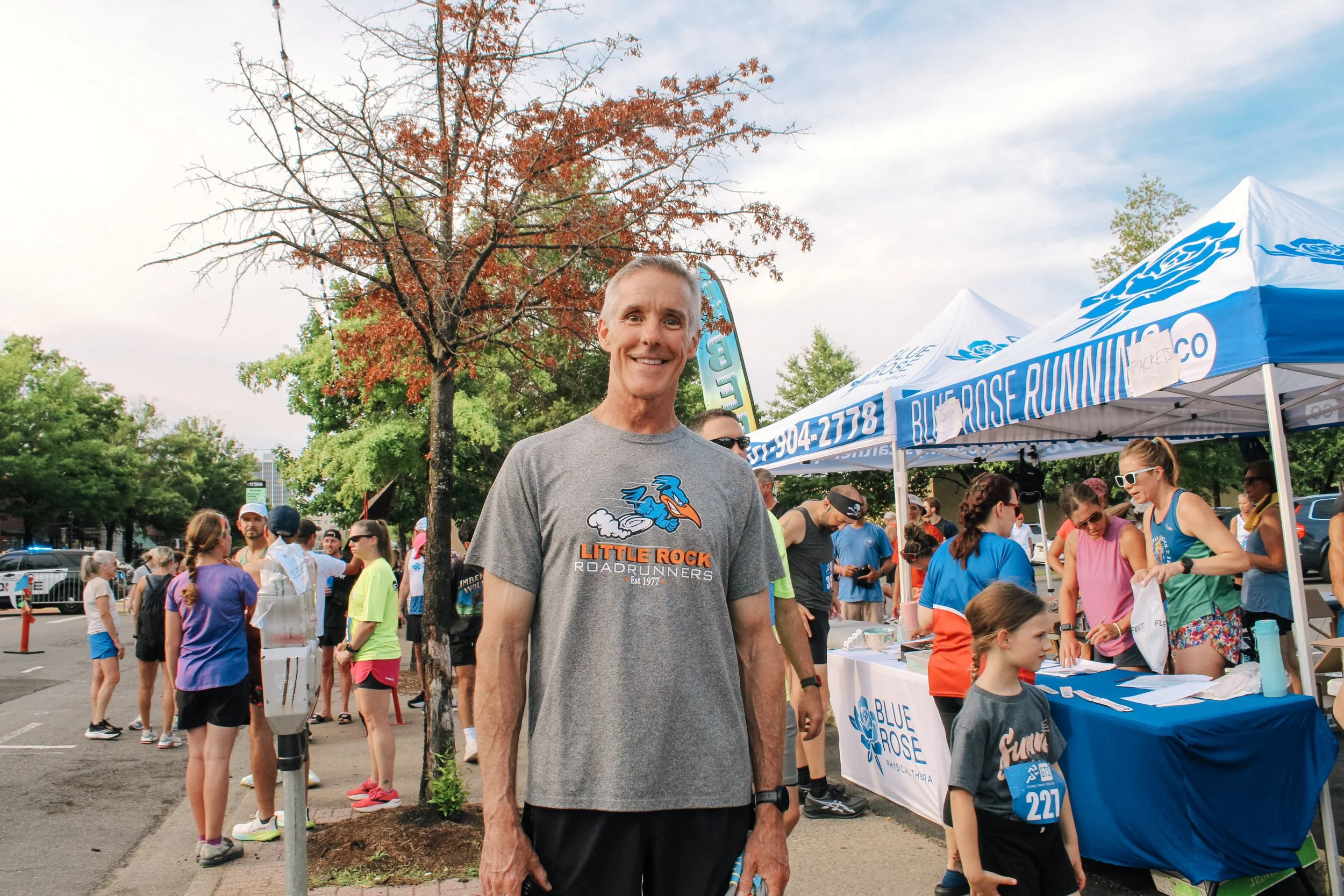 Man smiling at a running event wearing a gray T-shirt with a blue dragon logo and 'Little Rock Roadrunners' printed on it, surrounded by other participants and event booths with blue and white tents.