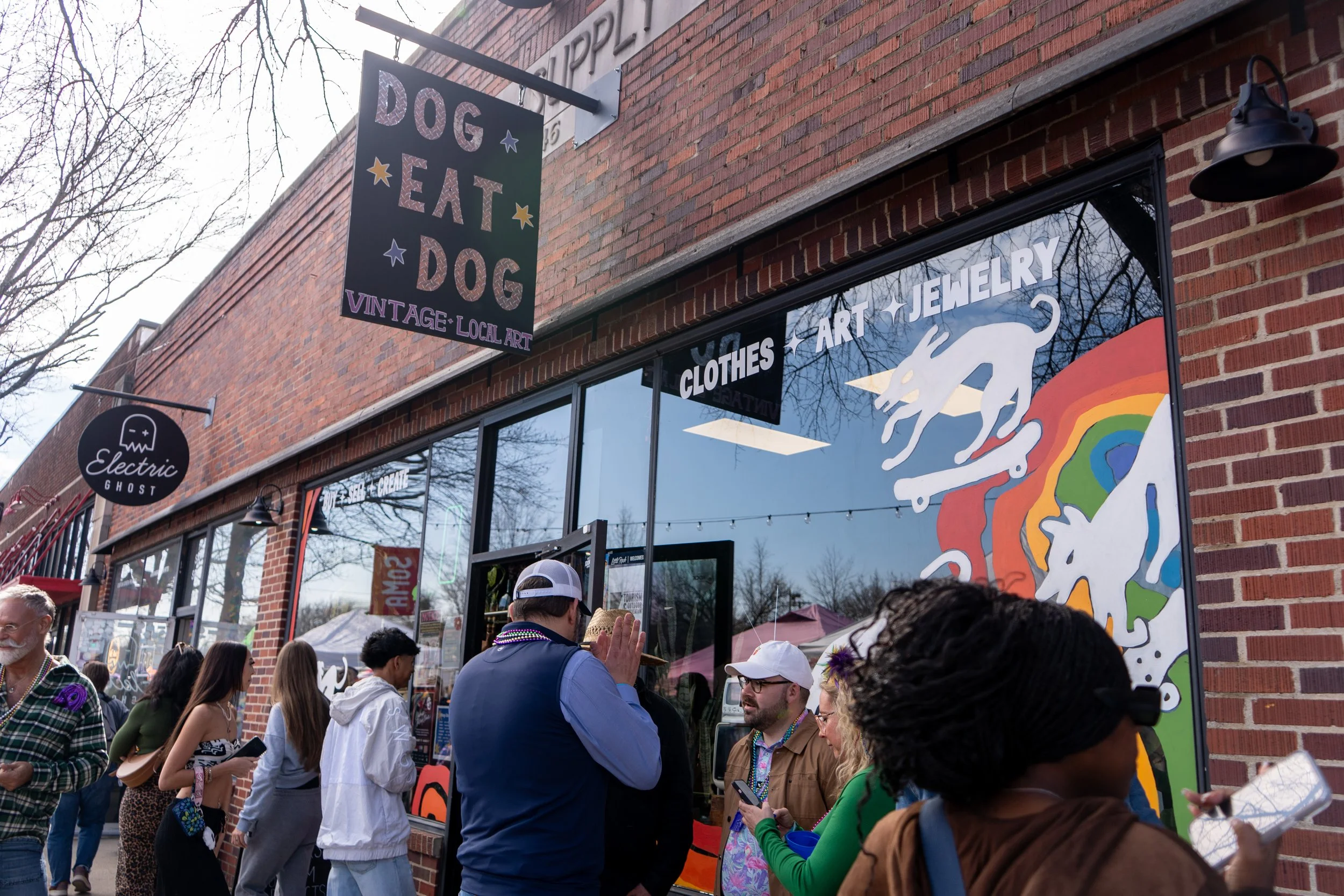 People standing outside a brick storefront with signs that say 'Dog Eat Dog' and 'Electric Ghost.' The window display features artistic designs including unicorns and a rainbow. Several individuals are engaged in conversation, some with phones.