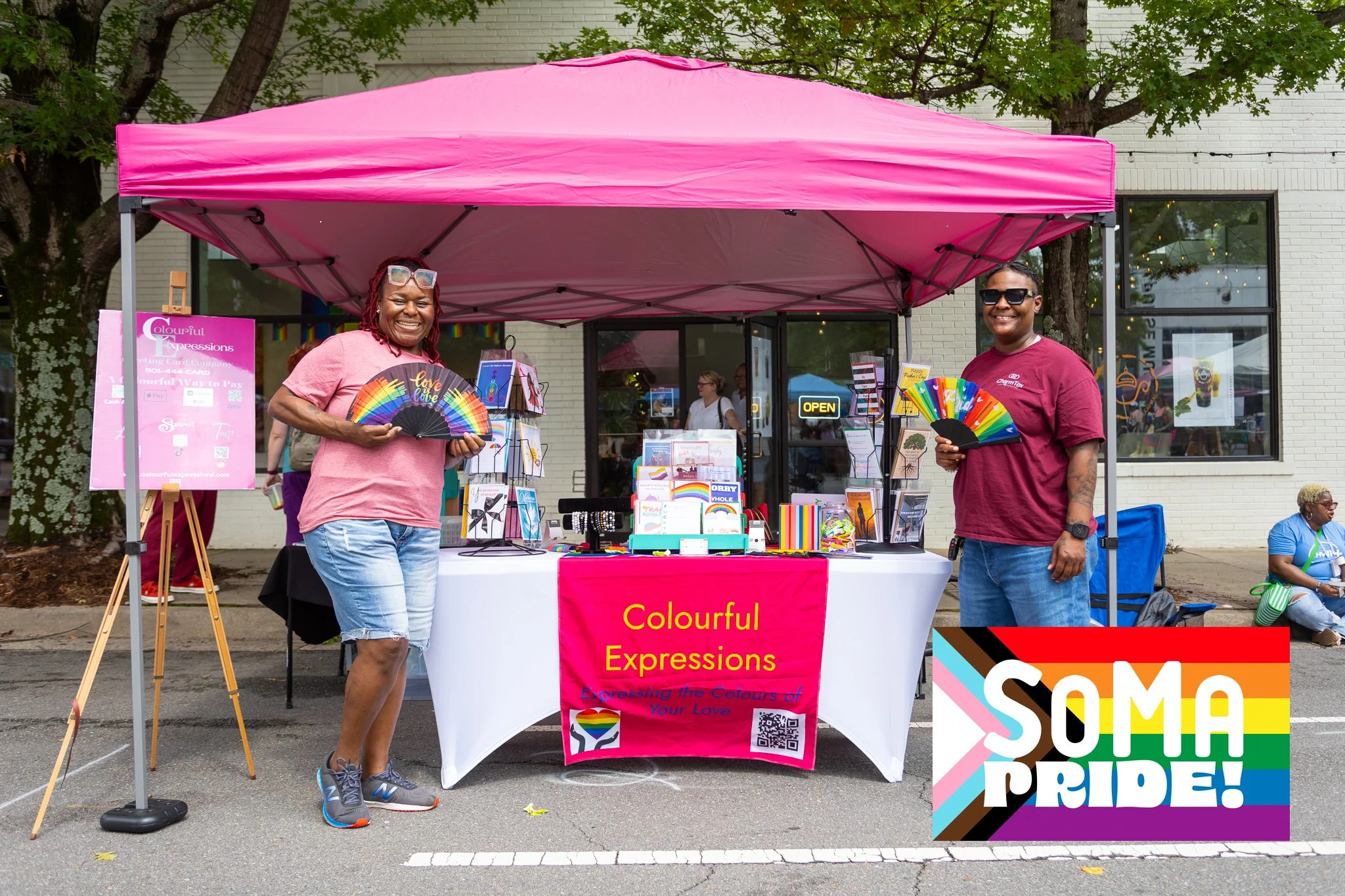 Two women smiling and holding rainbow-colored fans stand in front of a booth at an outdoor event, with a pink canopy, diverse merchandise on display, and a colorful 'SOMA PRIDE!' sign on the ground.