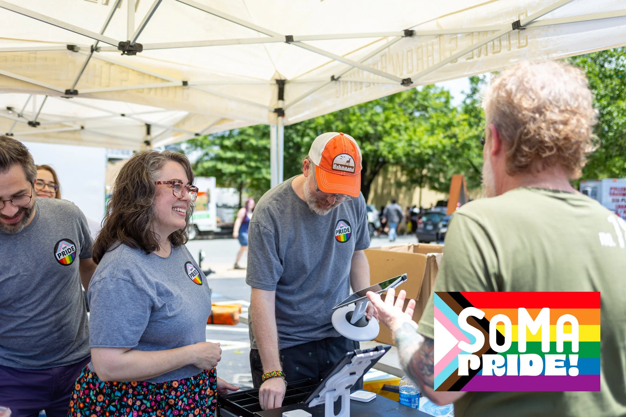 People at an LGBTQ+ Pride event, smiling and talking under a white canopy, with trees and other people in the background, and a colorful Pride logo in the corner.