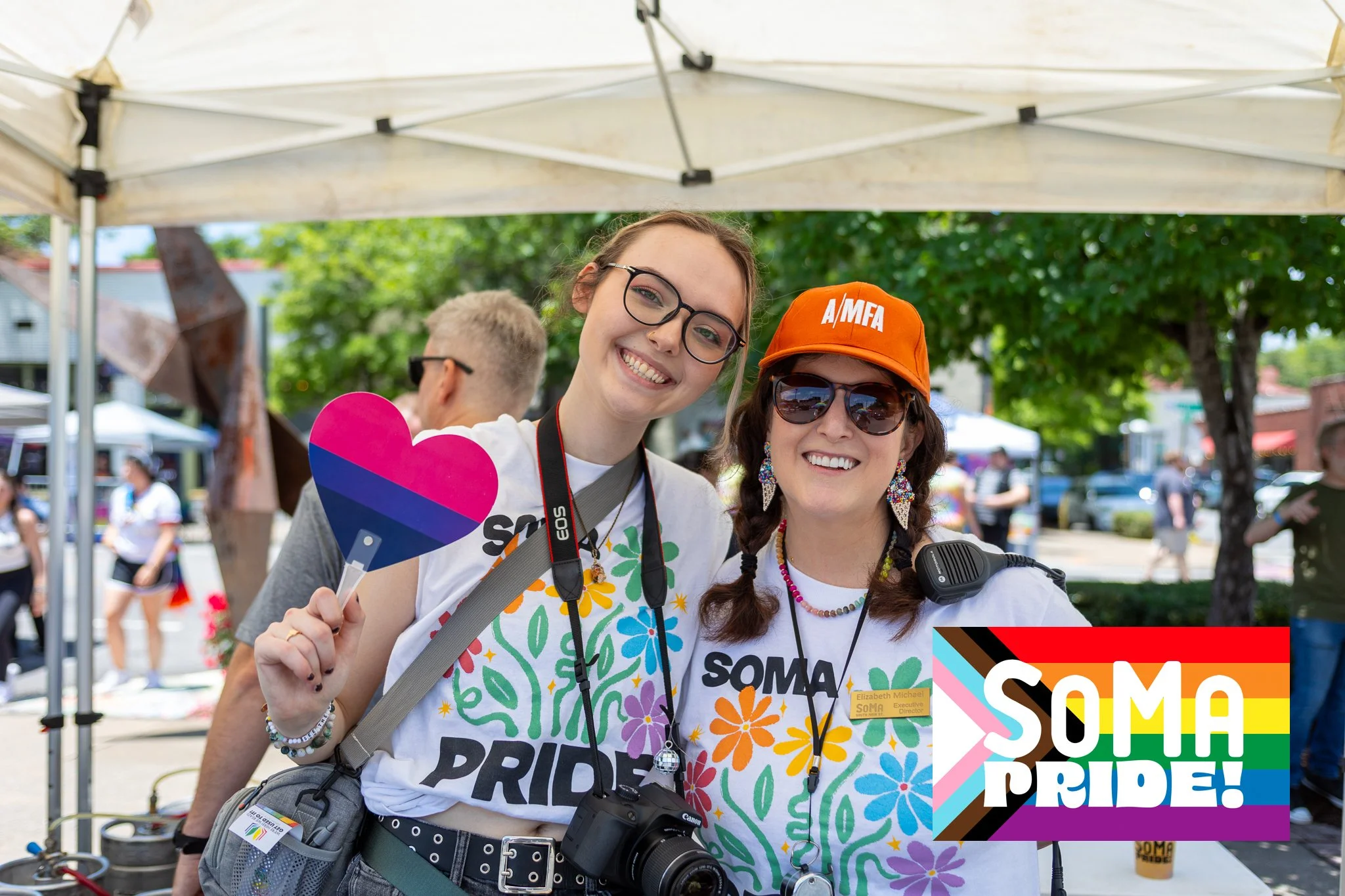 Two women smiling at a pride event under a white canopy. One woman has light skin, glasses, a white t-shirt with colorful floral designs, and is holding a pink, purple, and blue pride flag paddle. The other woman has light skin, dark hair in braids, glasses, an orange cap with white text, earrings, and a lanyard with a badge. They are standing outdoors with trees and people in the background, and a rainbow pride flag with the words "SOMA PRIDE!" is in the lower right corner.