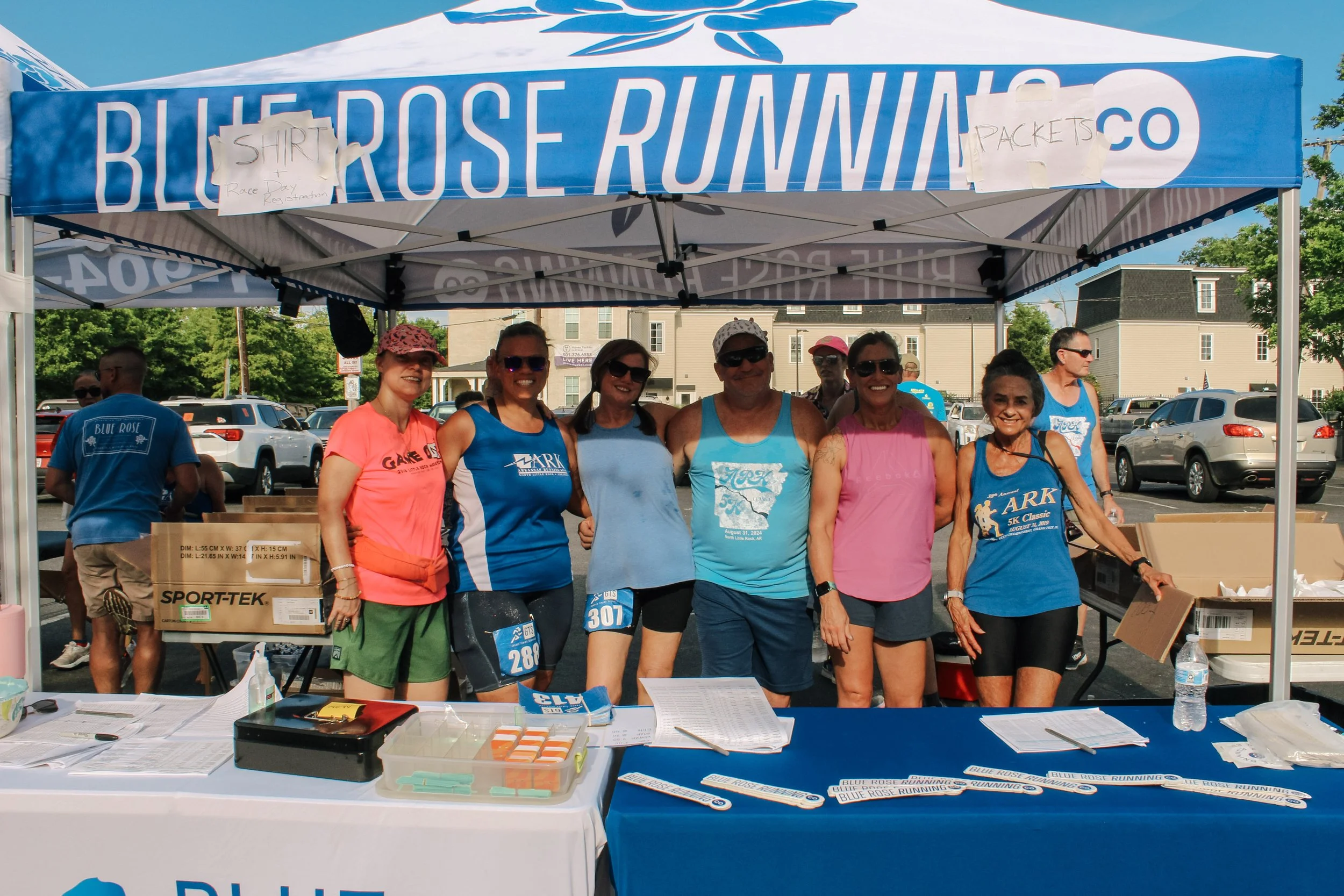 Group of people standing under a blue and white canopy at a race event, with registration tables and parked cars in the background.