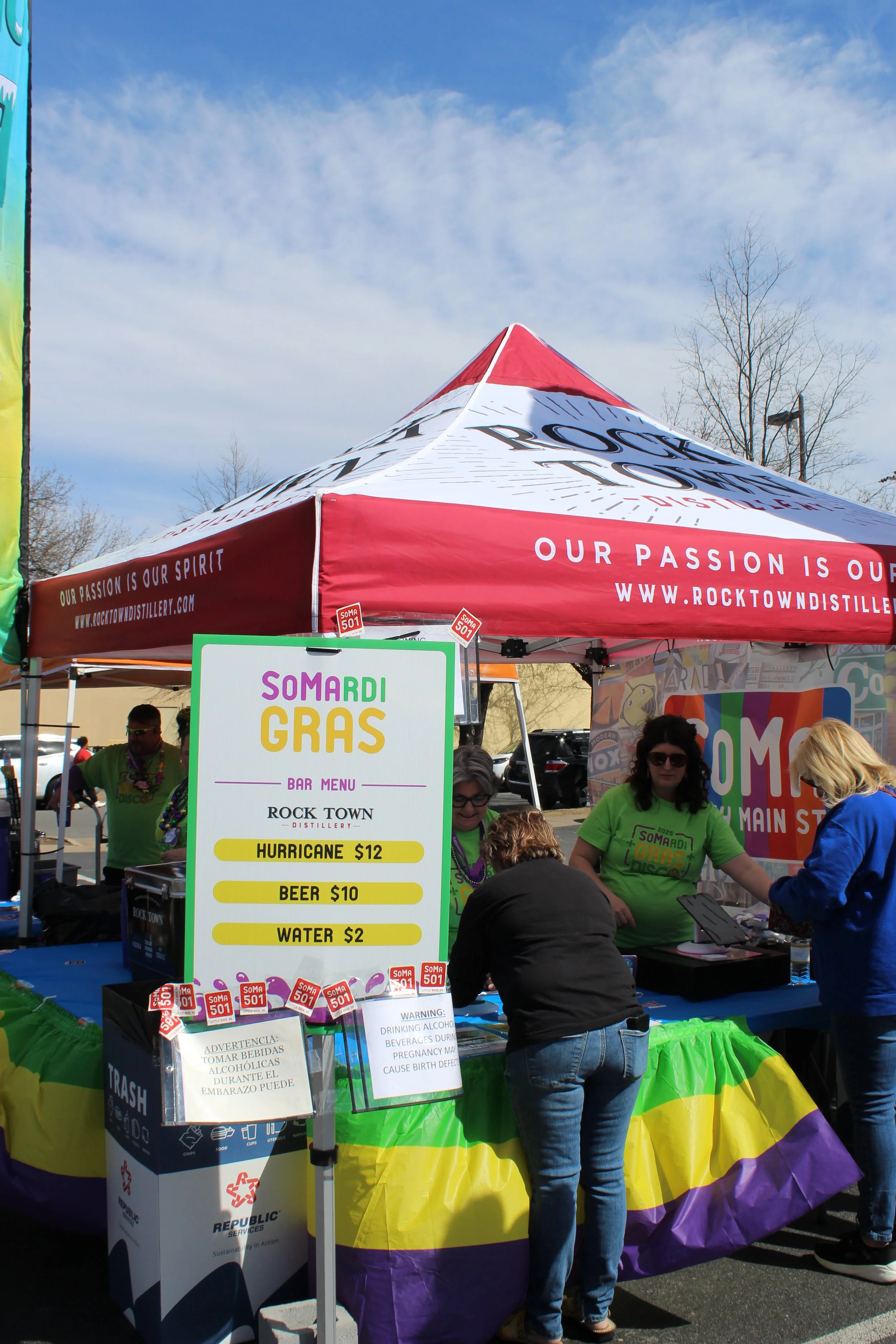 A booth at an outdoor event with a red canopy tent that reads 'Our passion is our spirit' and a sign listing food and drink prices for 'SOMARi Gras' at Rock Town Distillery. People are ordering at the booth, which is decorated with rainbow colors.