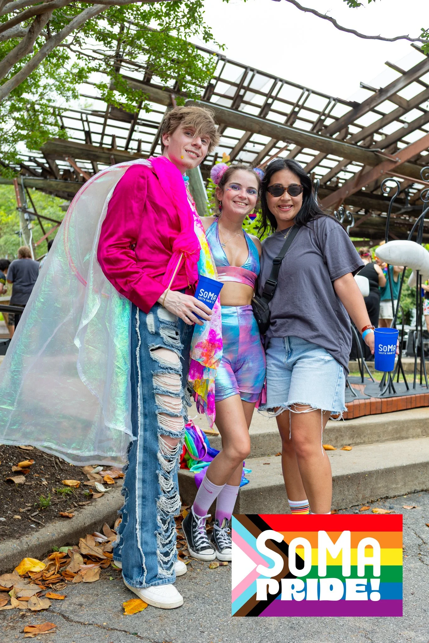 Three people smiling and posing outdoors at a pride event, with a wooden structure and trees in the background, and a colorful ''SOMA Pride'' sign at the bottom right corner.