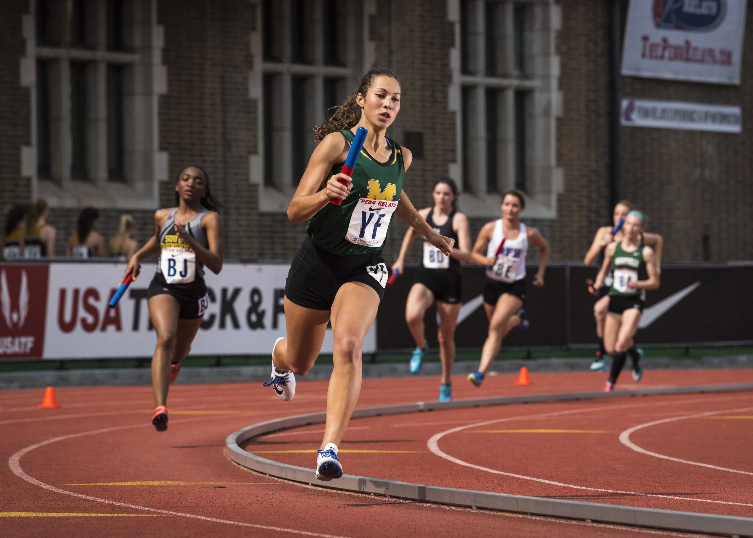 Female athletes competing in a relay race on a red track at night, with some runners holding relay batons and a banner that reads USA Track & Field in the background.