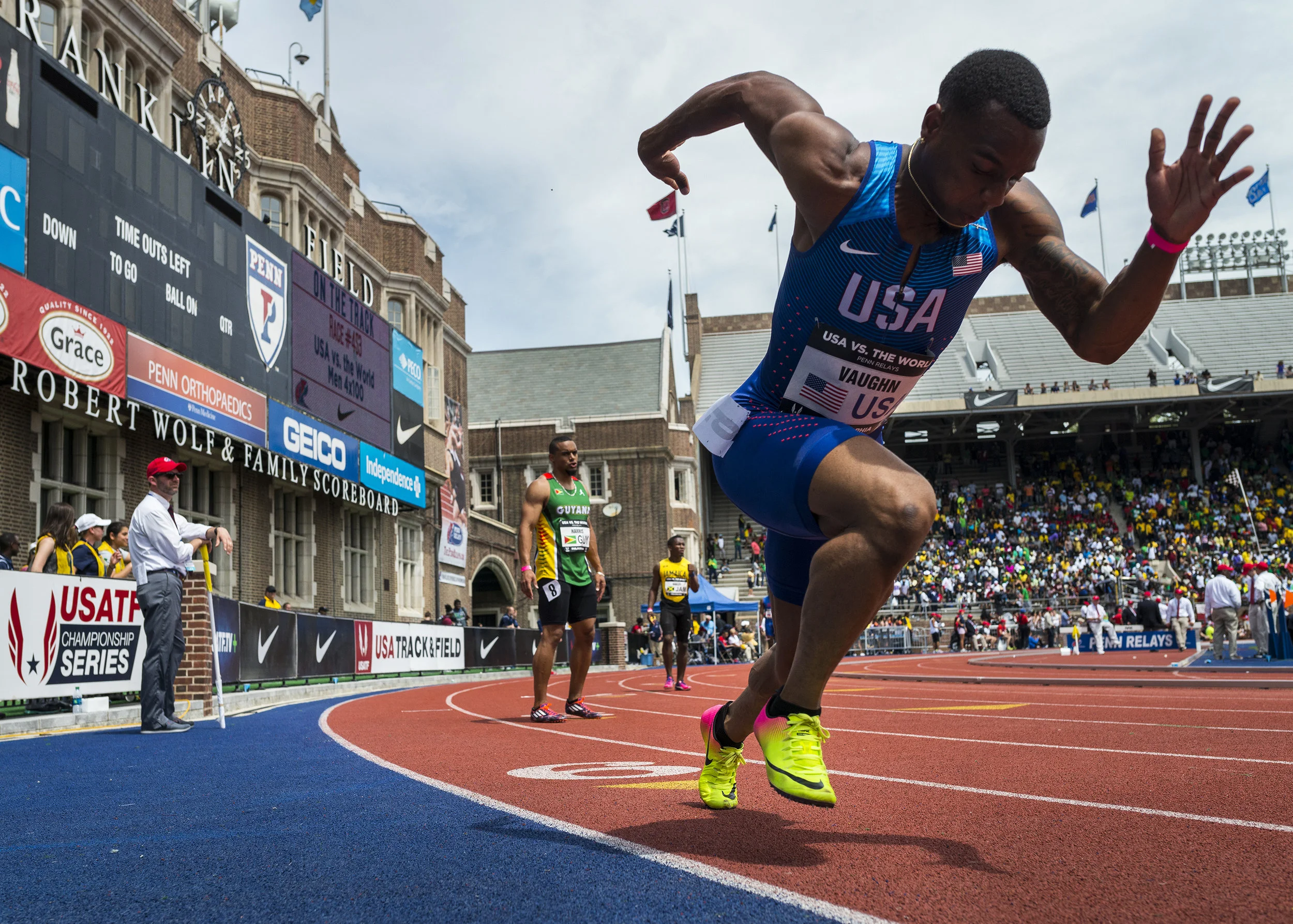 American male sprinter in blue USA uniform sprinting on a red track during a race at a stadium with spectators in the stands.