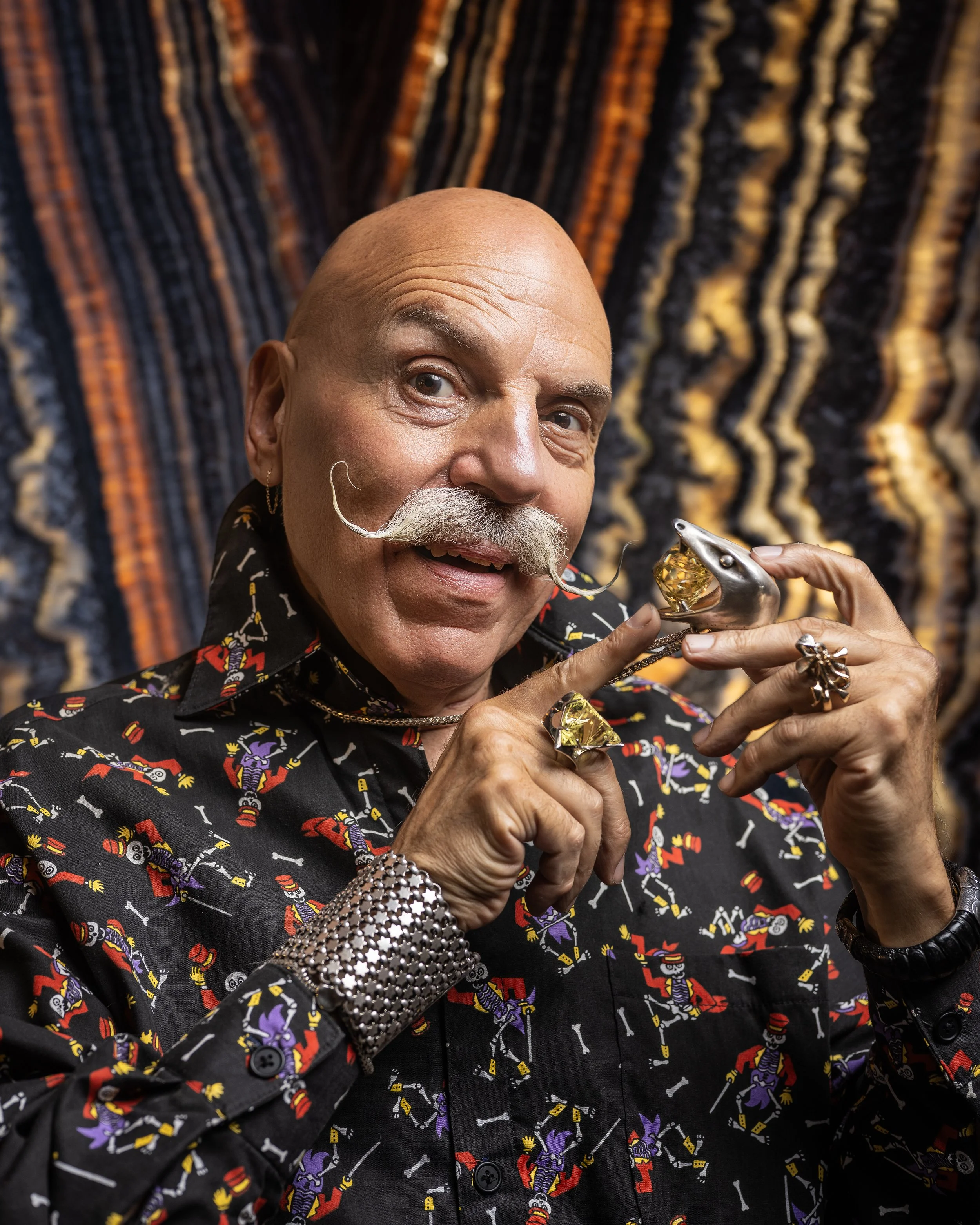 A man with a bald head and a mustache holding a silver dolphin-shaped object and wearing colorful jewelry, standing in front of a patterned background.