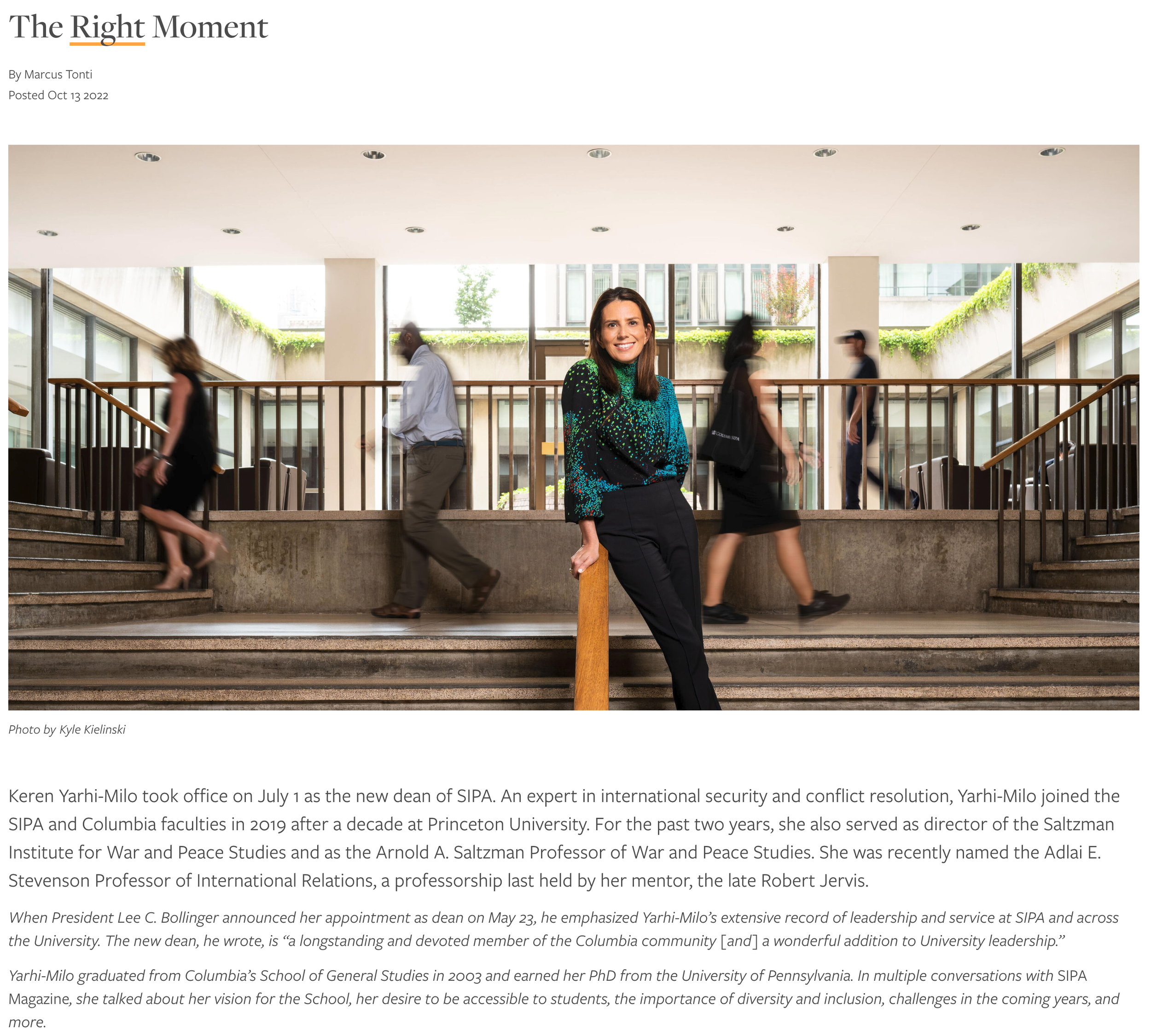 Columbia University SIPA Dean. A woman standing on stairs, leaning on a railing in a modern building with people walking behind her. She is smiling, wearing a colorful blouse and black pants.
