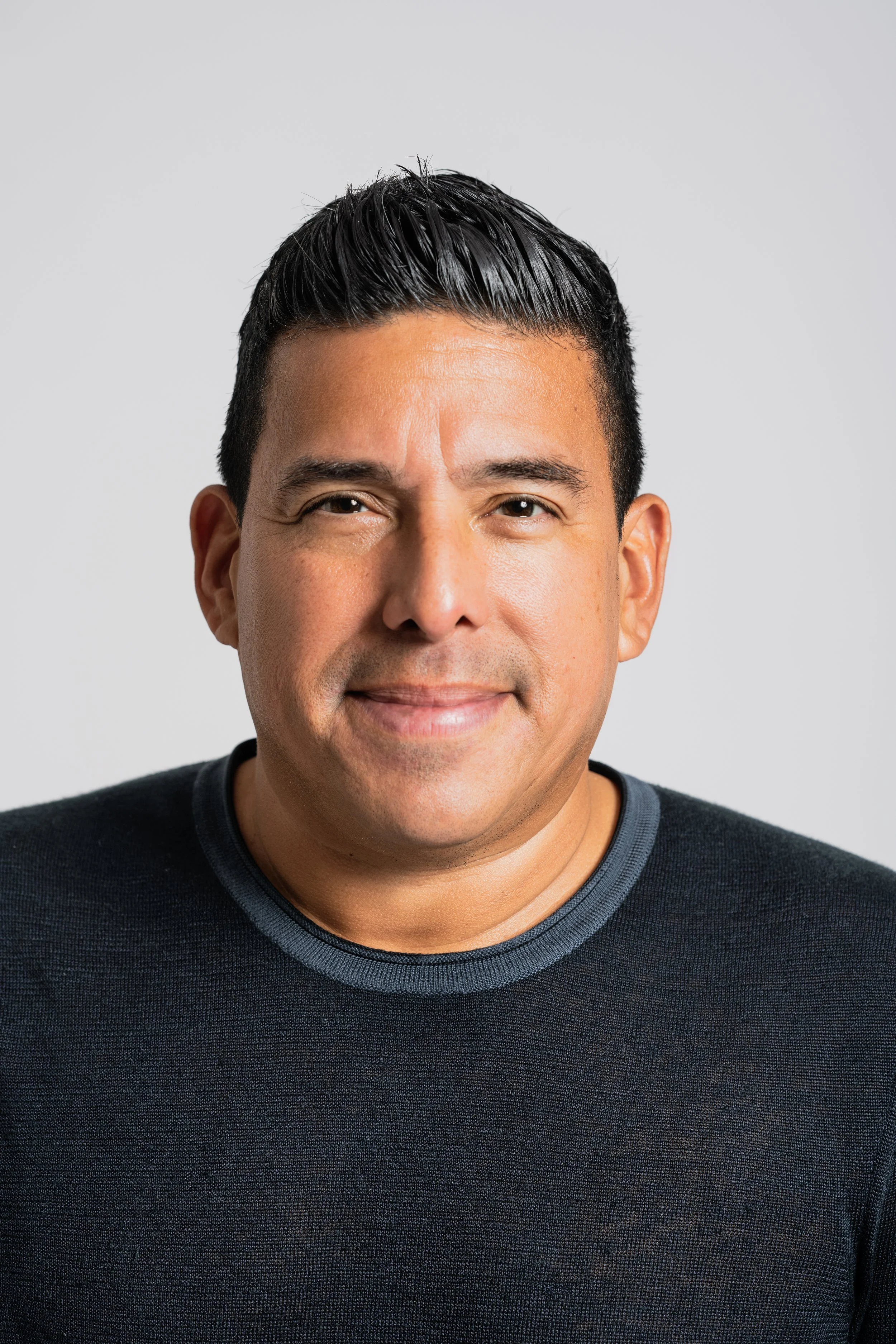 Close-up of a smiling man with dark hair, wearing a black sweater with a gray collar, standing in front of a plain light gray background.