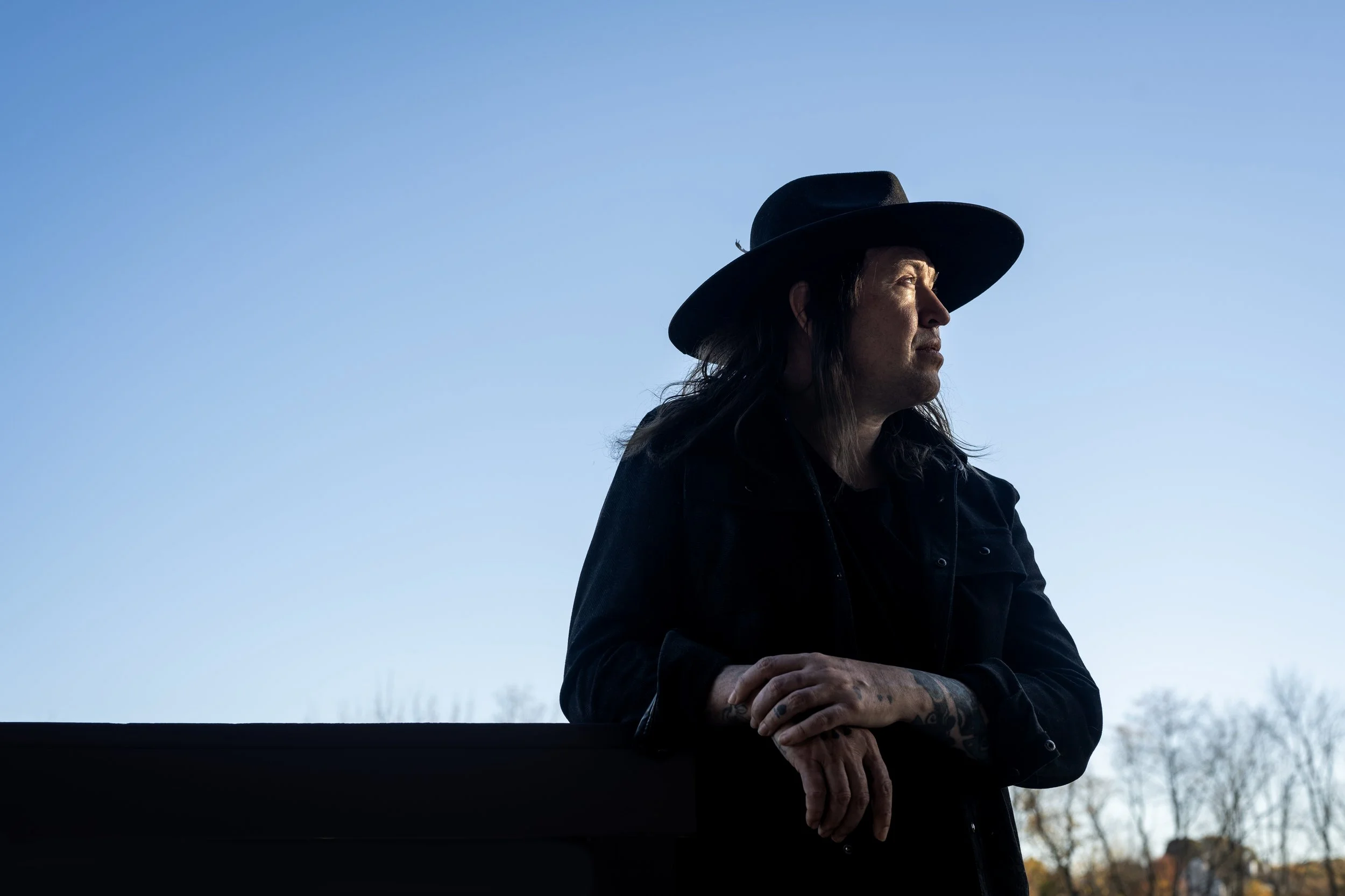 A person with long dark hair, tattoos on their hand, wearing a black hat and jacket, looking to the right against a clear blue sky with some trees in the distant background.