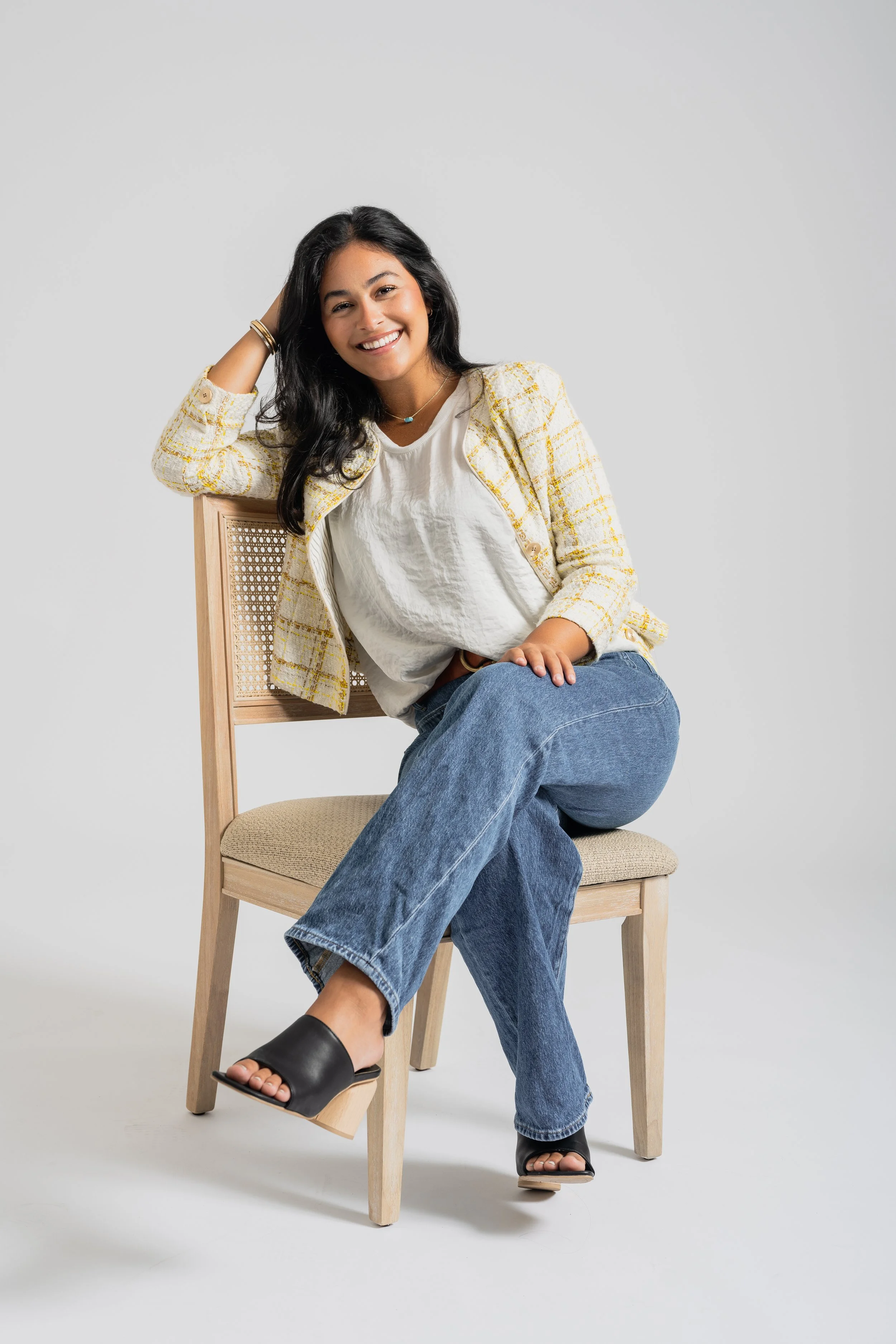 Smiling woman in white t-shirt and yellow plaid blazer sitting on a wooden chair with crossed legs, black open-toe heels, and dark hair, posing against a plain gray background.