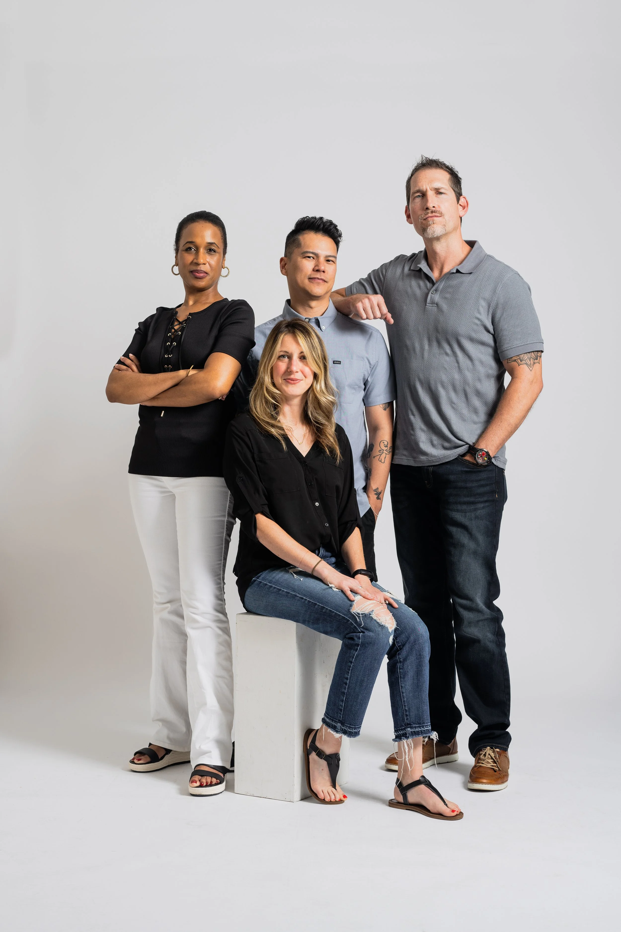 Group of five diverse people posing together against a plain white background, with one person sitting on a stool and four standing.