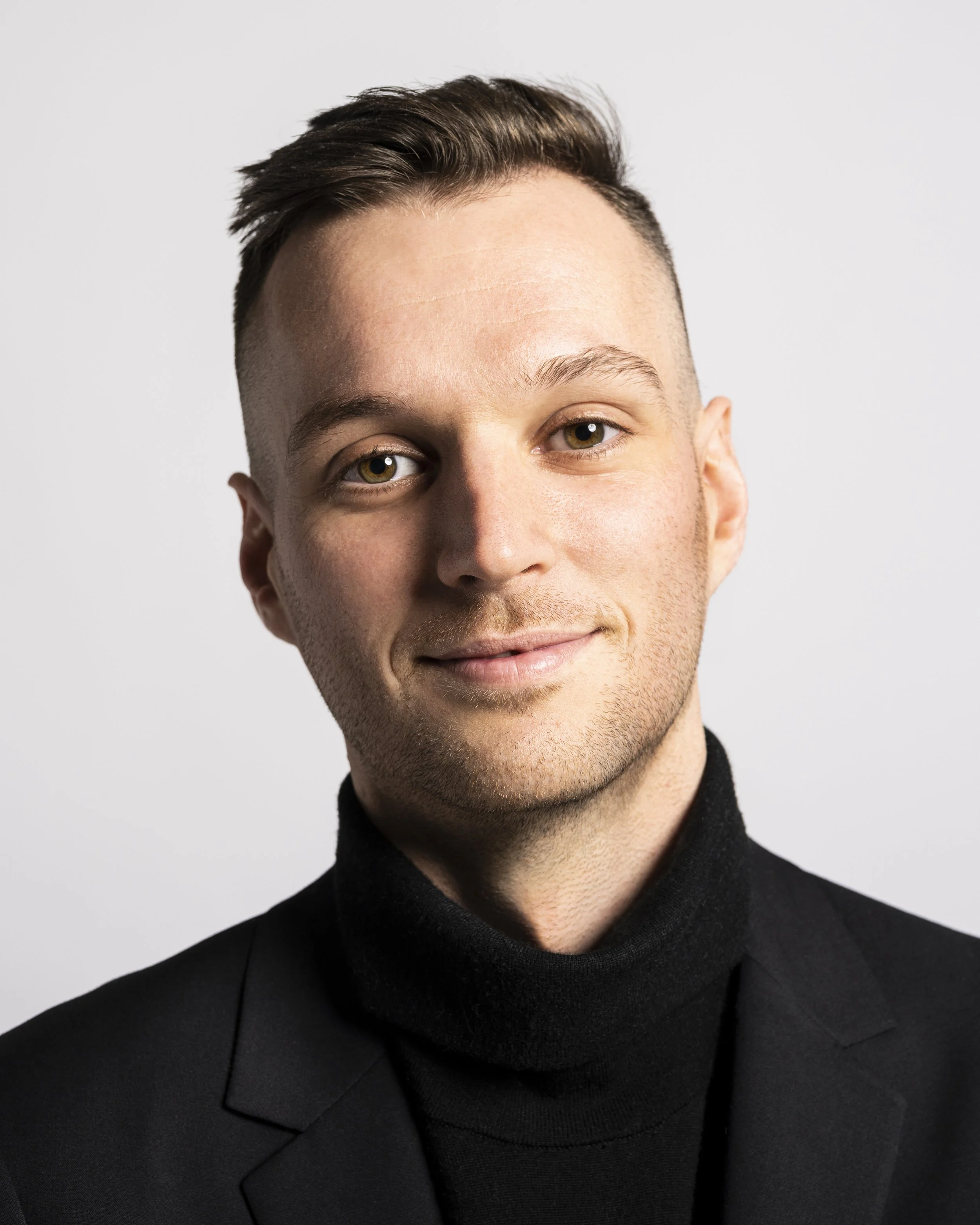 A portrait of a young man with light skin, short brown hair, wearing a black turtleneck and blazer, smiling slightly against a plain white background.