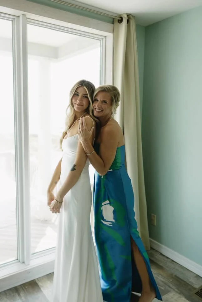 Two women standing together, one in a white dress and the other in a colorful dress, inside a room with light green walls and a sliding glass door with curtains.