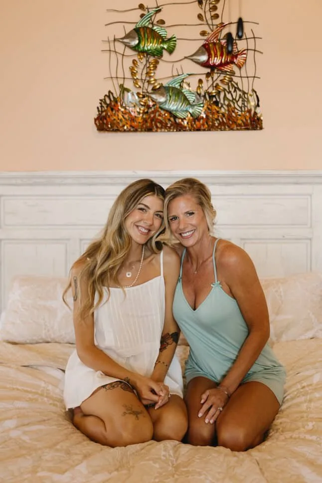 Two women sitting on a bed, smiling, with colorful fish wall art above them.