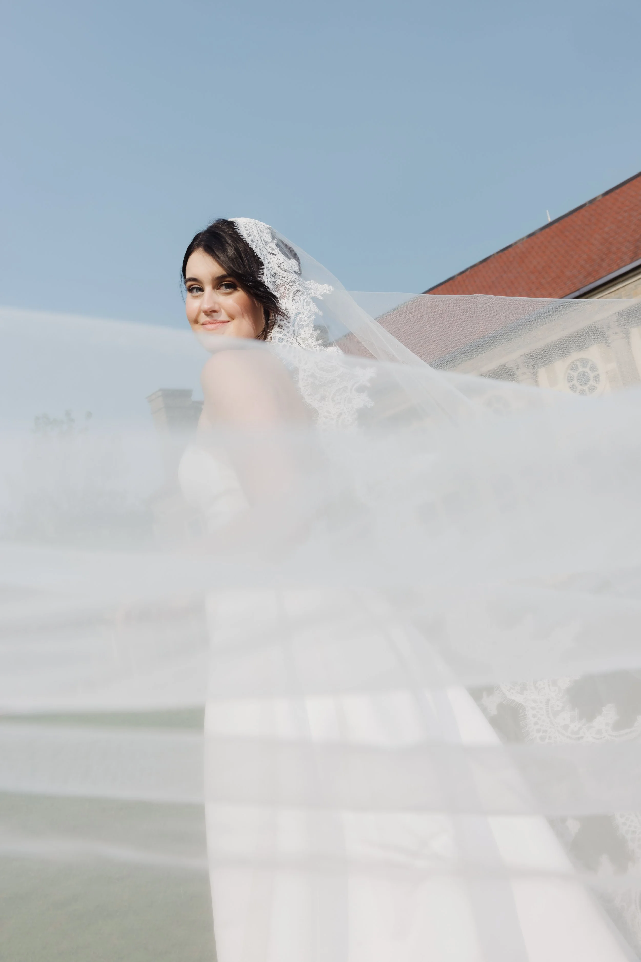 A bride with dark hair and blue eyes, wearing a white lace wedding dress and veil, smiling outdoors with a building and blue sky in the background.