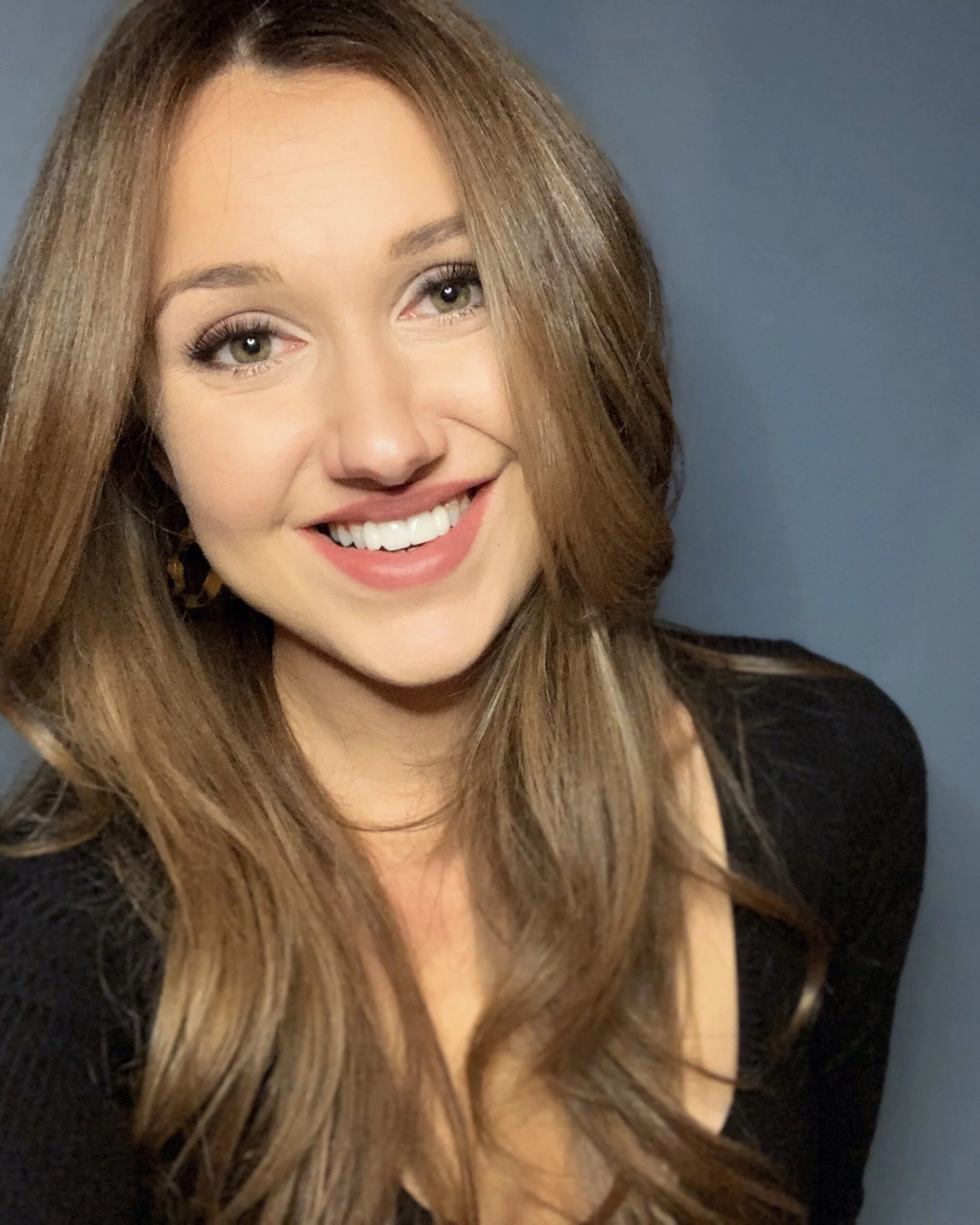 A woman with long, wavy, light brown hair, smiling, wearing a black top, standing against a plain blue background.