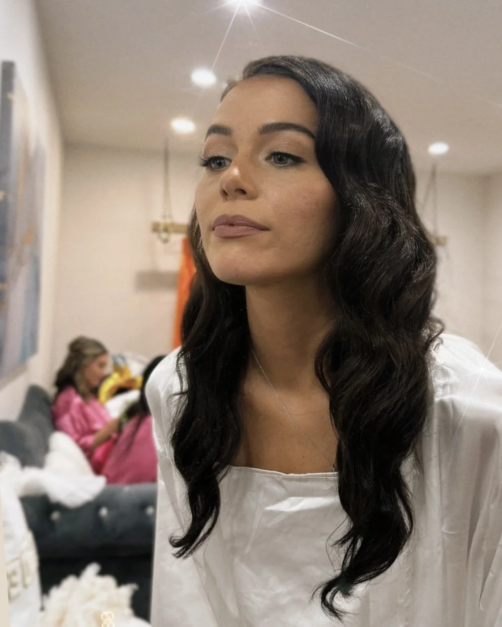 A woman with long, dark, wavy hair taking a selfie in an indoor setting. In the background, a girl in a pink outfit is sitting on a sofa, possibly on a phone or tablet.