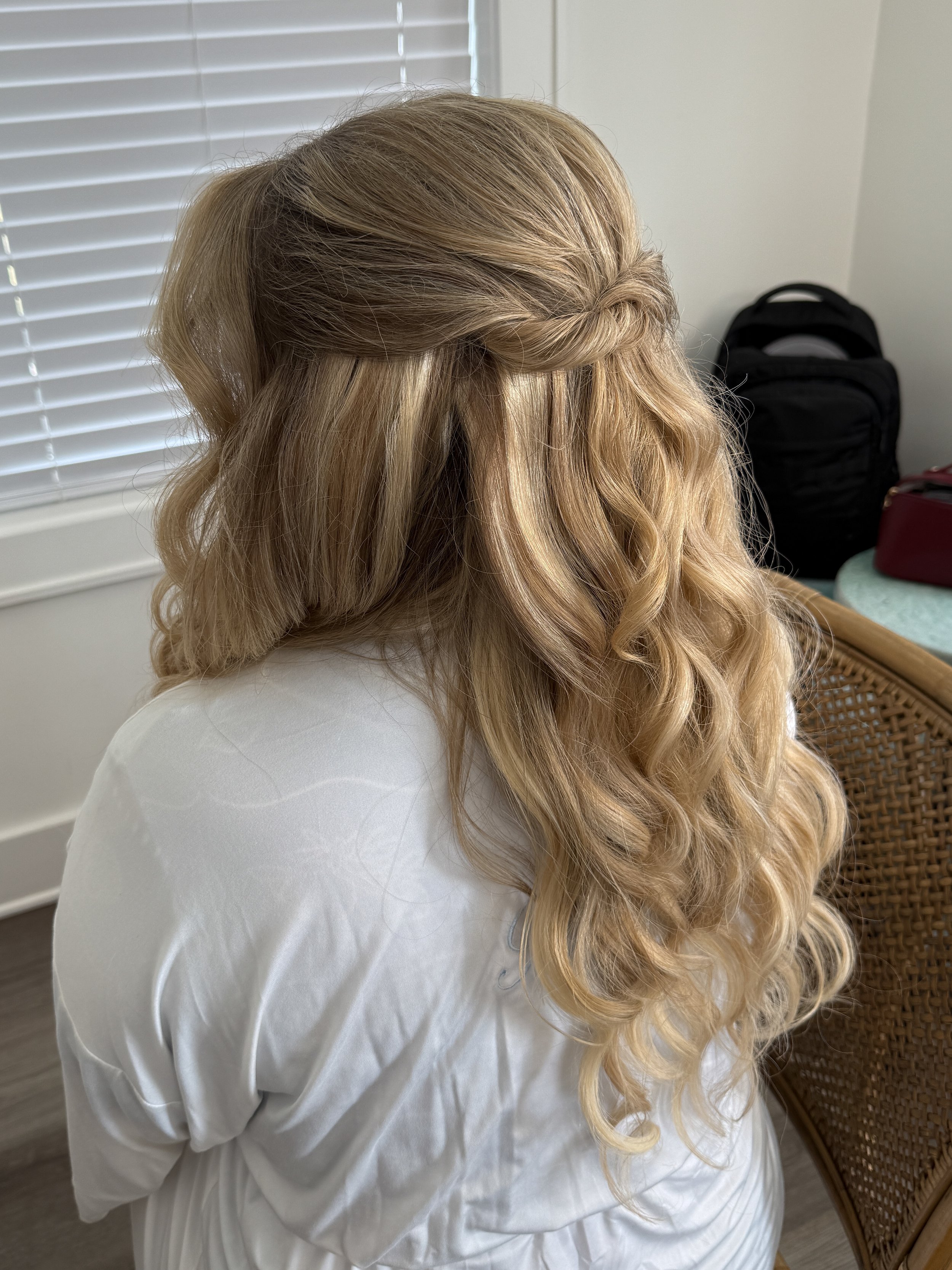 A woman with long, blonde, curly hair styled in loose waves sitting on a wicker chair with a white shirt on, next to a window with closed blinds.