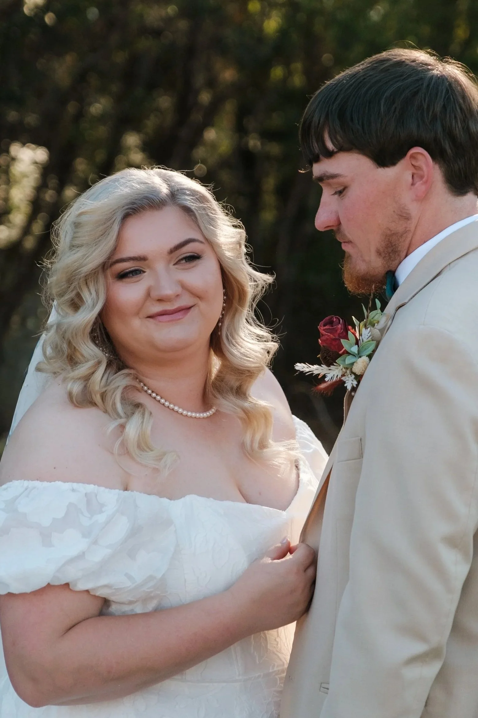A bride and groom standing outdoors during their wedding, holding hands, with trees in the background.