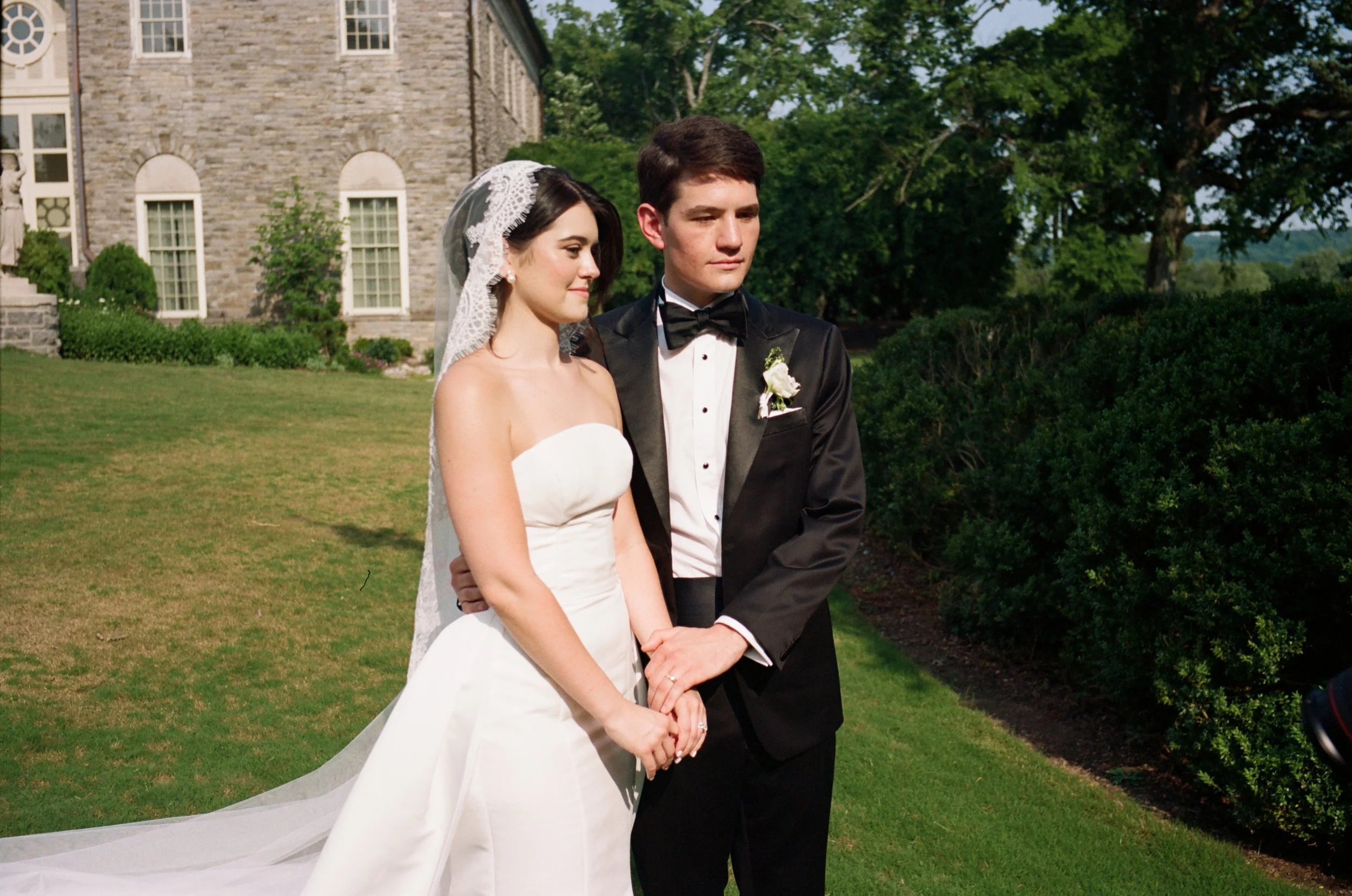 A bride and groom standing outside during their wedding, holding hands and looking contemplative. The bride is wearing a strapless white gown with a lace veil, and the groom is dressed in a black tuxedo with a bow tie.