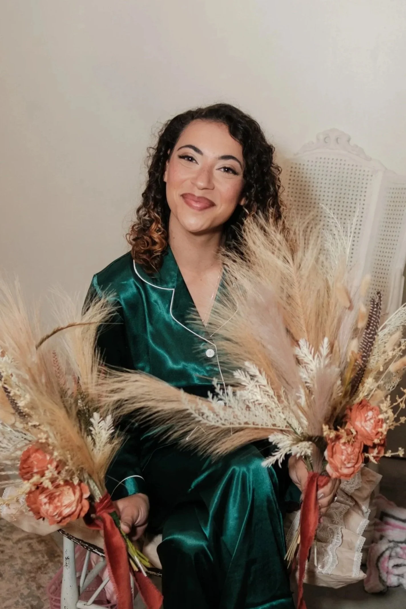 A young woman with curly hair and a green satin pajama top sitting in a chair, smiling, surrounded by a large bouquet of dried flowers and pampas grass.