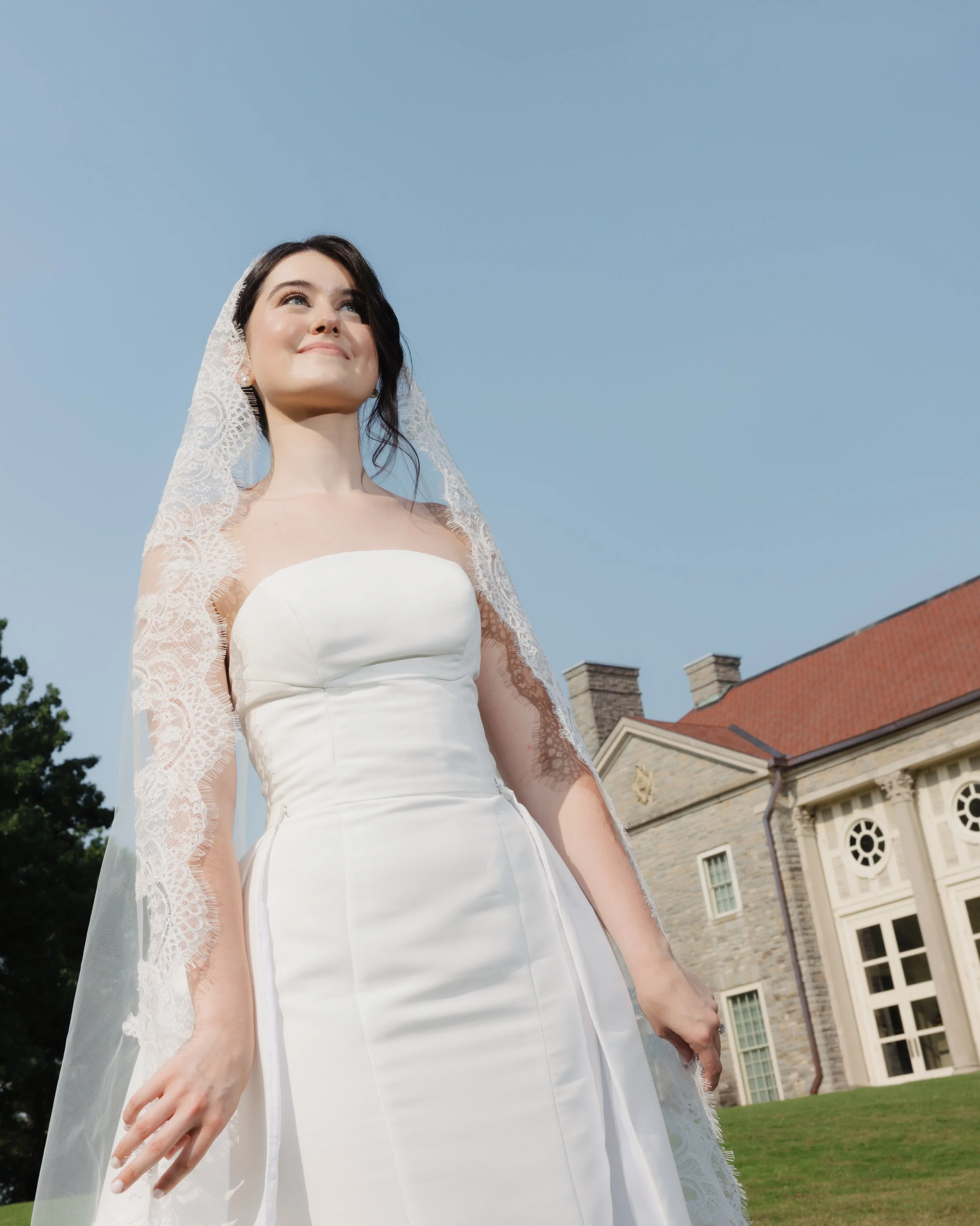 A bride in a white wedding dress and lace veil standing outside in front of a historic building with red roof and large windows under a clear blue sky.