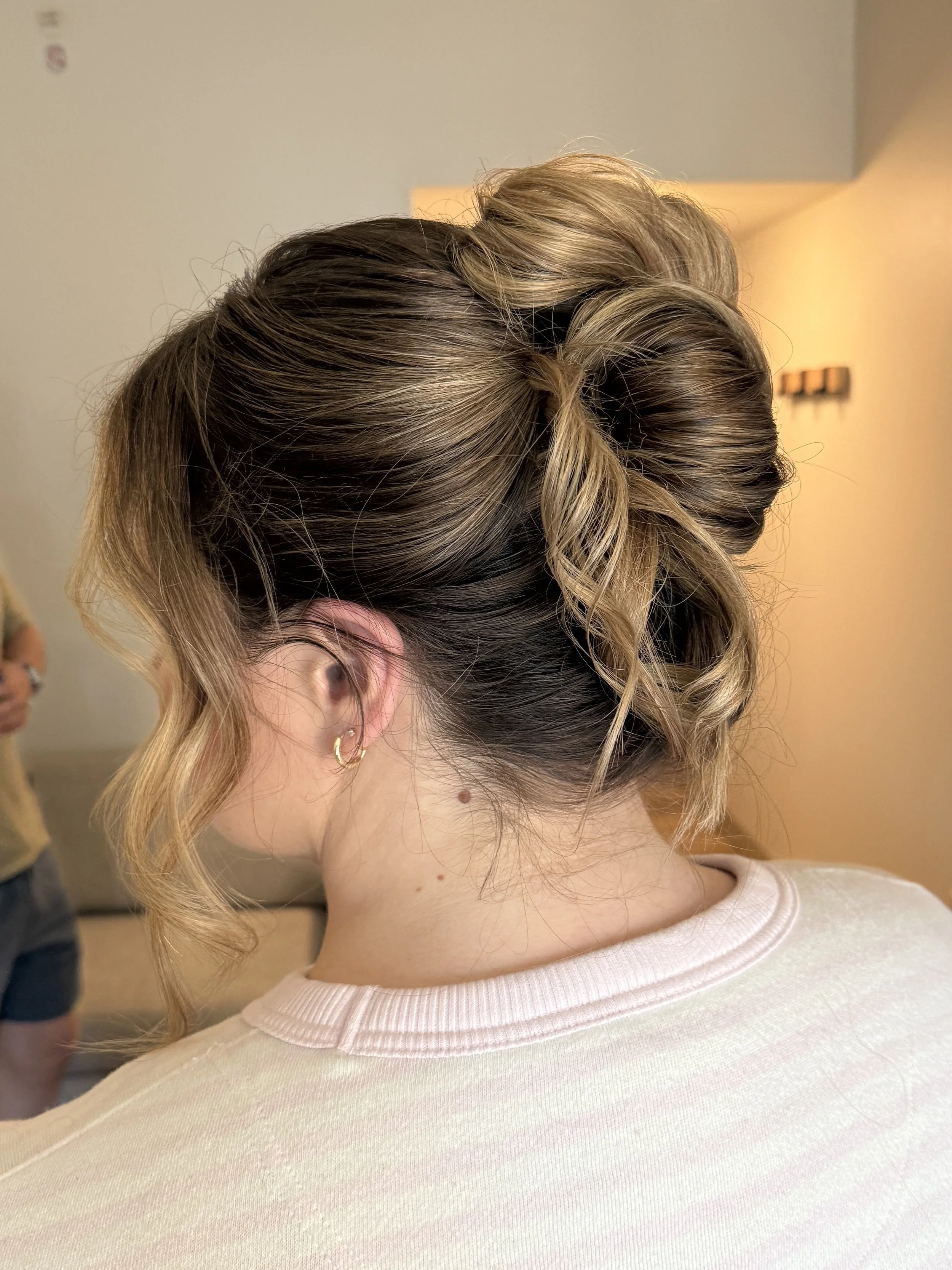 Close-up of a woman's hairstyle with blonde and brown wavy hair styled in an elegant updo with loose curls, worn with small hoop earrings, seen from behind.