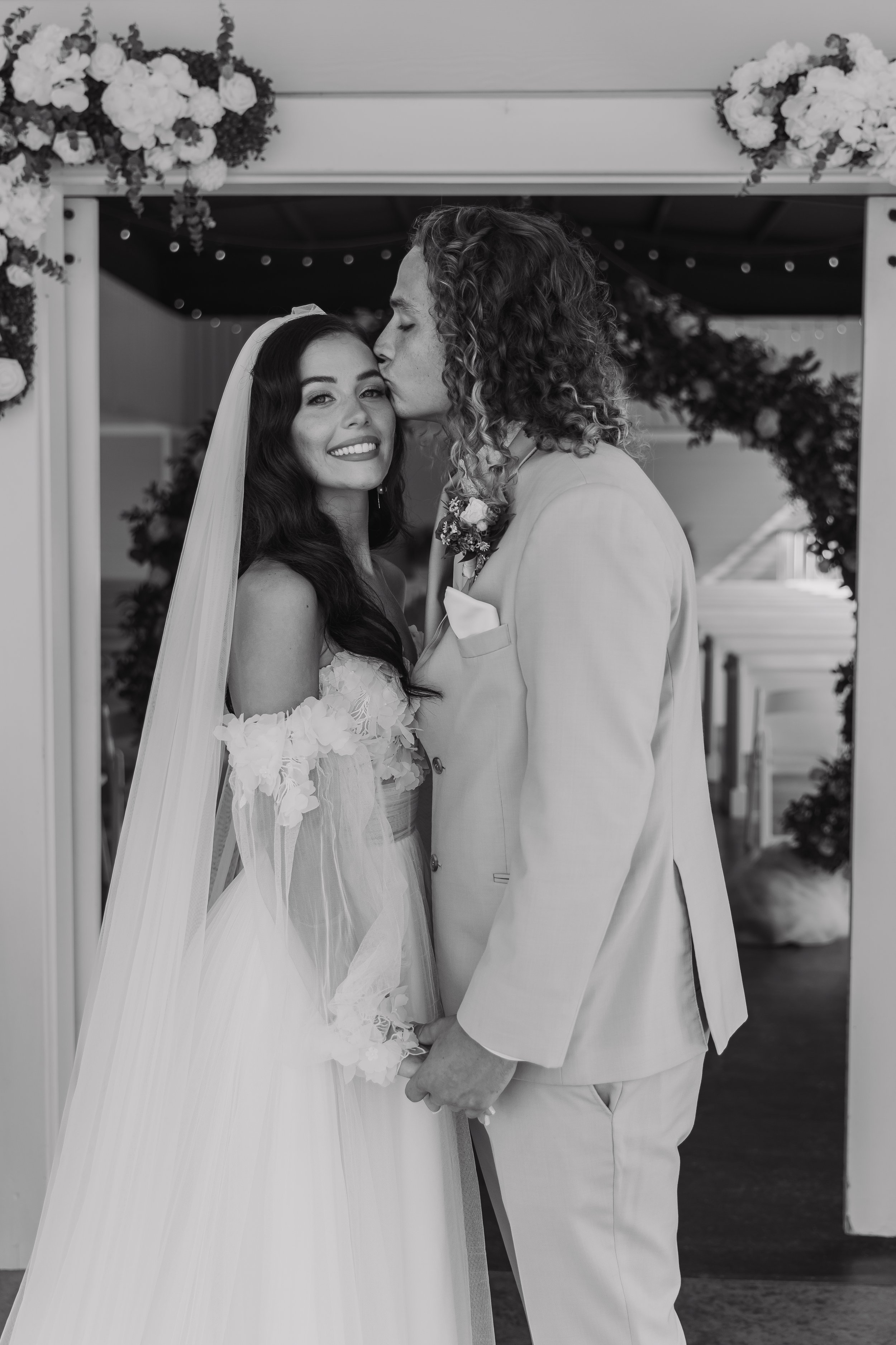 Black and white photo of a bride and groom at their wedding, holding hands under an arch decorated with flowers, with the groom kissing the bride's forehead.