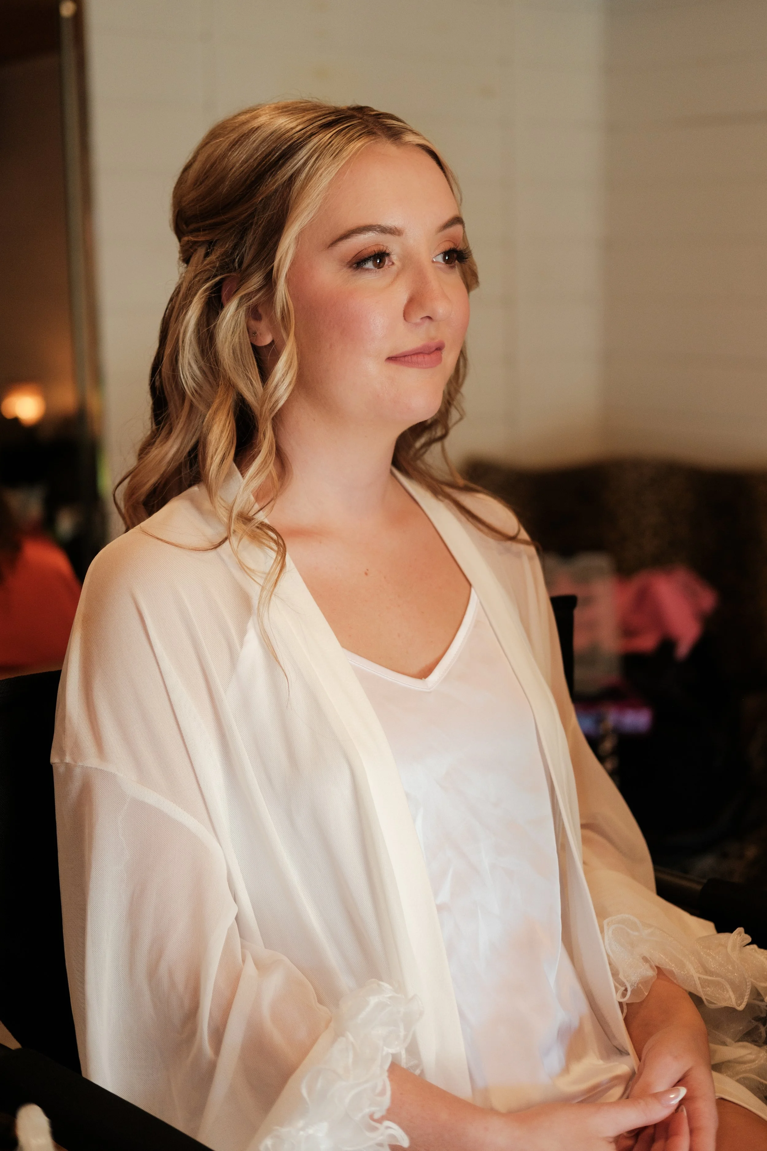 A woman with blonde, wavy hair sitting in a room, dressed in a white satin top and a sheer, cream-colored robe, gazing softly ahead.