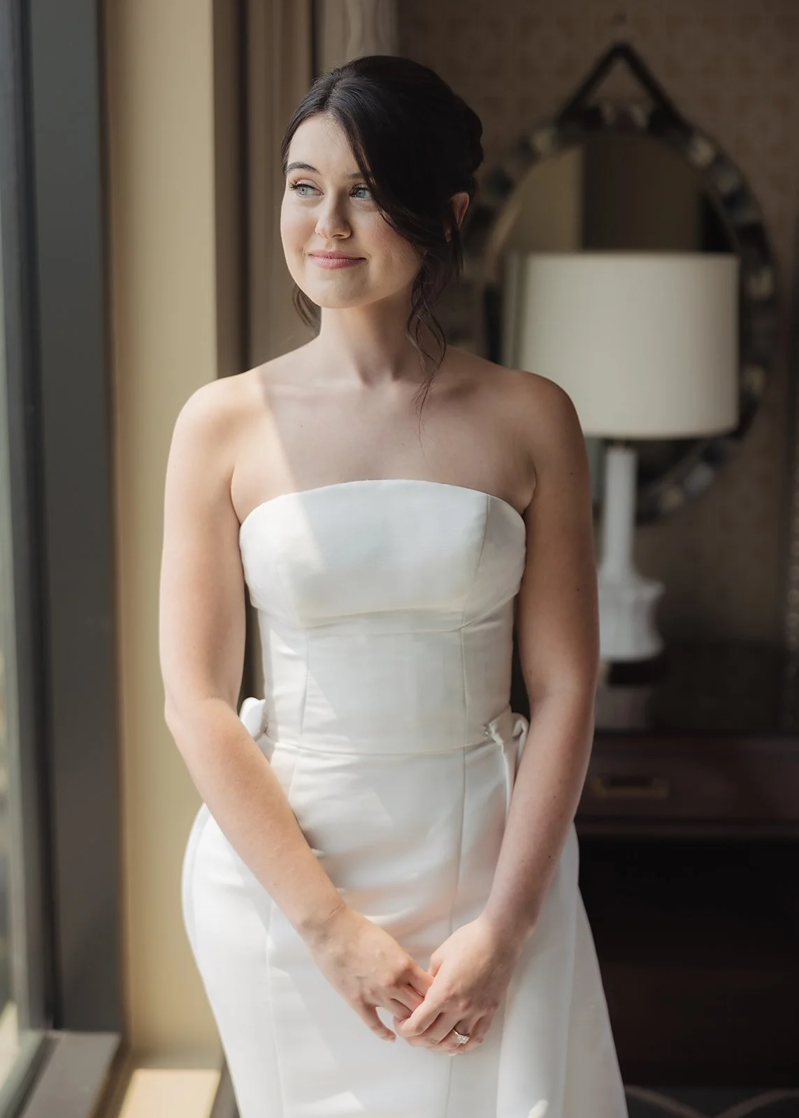 A woman in a white strapless wedding dress standing indoors, looking out a window with a gentle smile.
