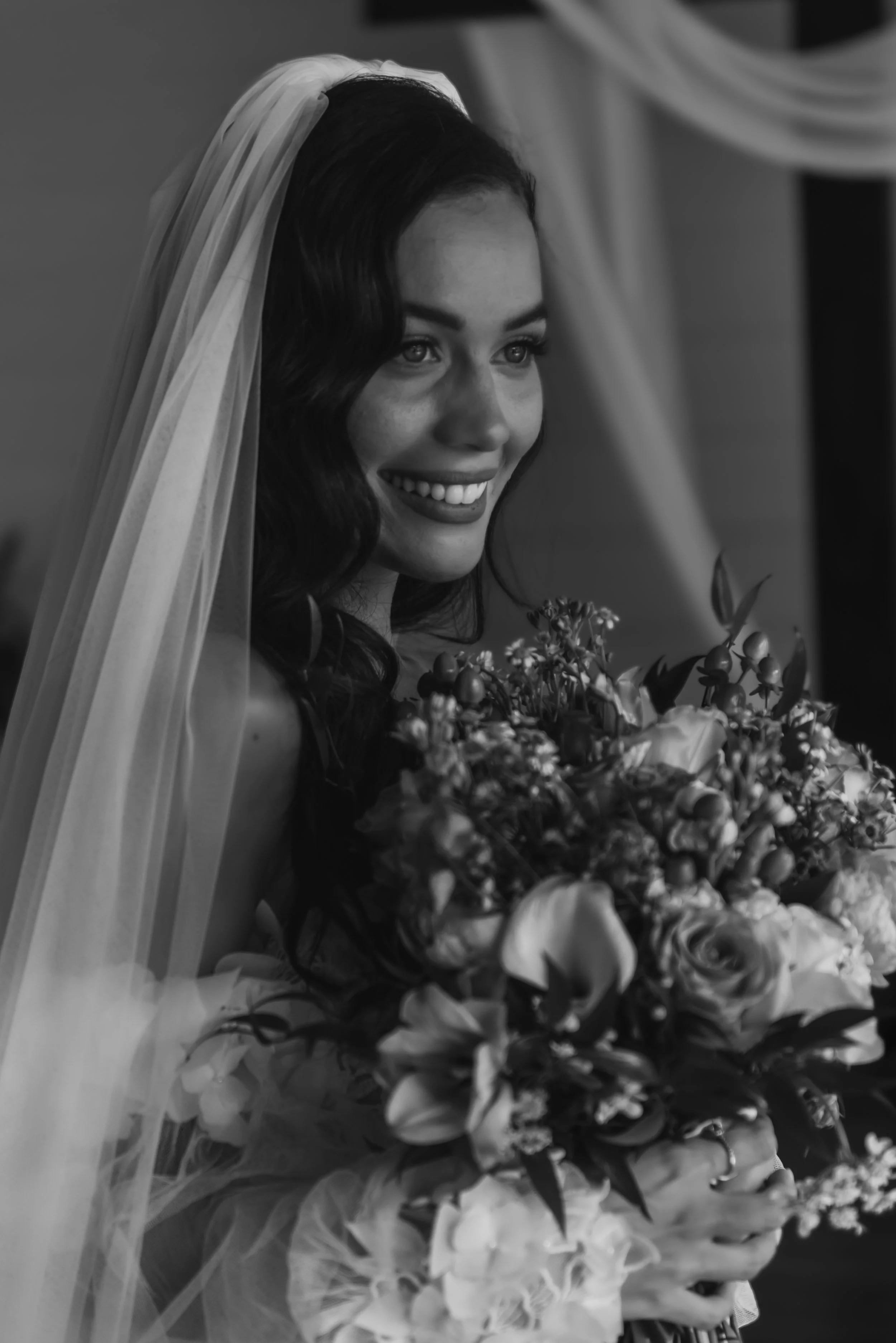 Black and white photograph of a bride smiling, wearing a veil and holding a bouquet of flowers.