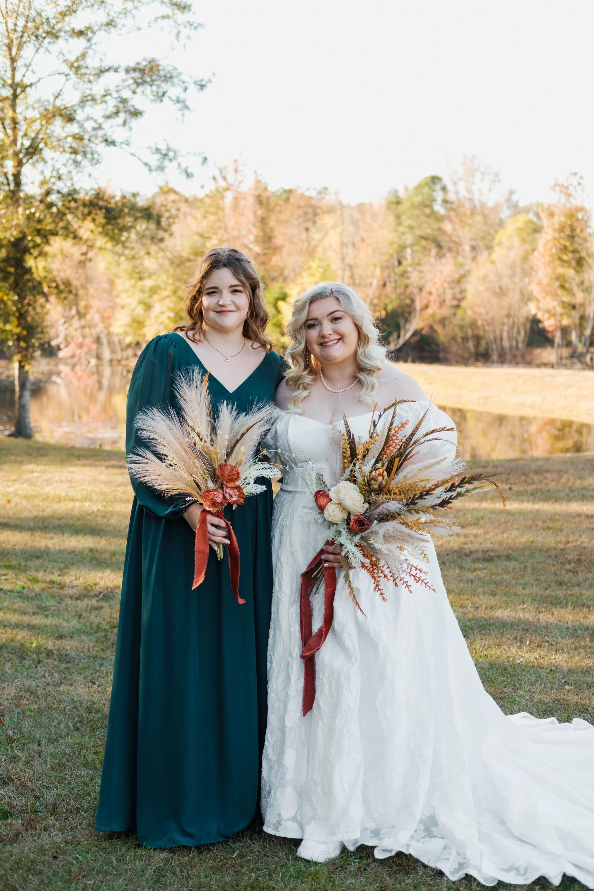 Two women standing outdoors in a park during autumn, holding bouquets of dried flowers and grasses, with trees and a lake in the background. One woman is wearing a white wedding dress, and the other is wearing a dark green dress.